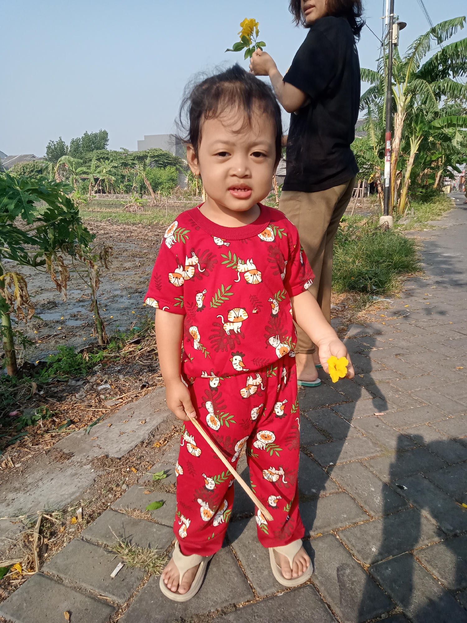 A young child, appearing to be around two to three years old, stands facing the camera outdoors. The child is wearing a red pajama set with a white pattern of cats and leaves, and light-colored sandals. They are holding a thin wooden stick in their right hand and a single yellow flower in their left hand. Behind the child, a person wearing a black t-shirt and brown pants stands with their back to the camera, holding a small bouquet of yellow flowers. The background features a street with a paved walkway, grass, and trees, including banana trees. The ground appears to be uneven and muddy in some areas. The sky is clear and blue, suggesting a sunny day. The location is identified as Unknown, Unknown.