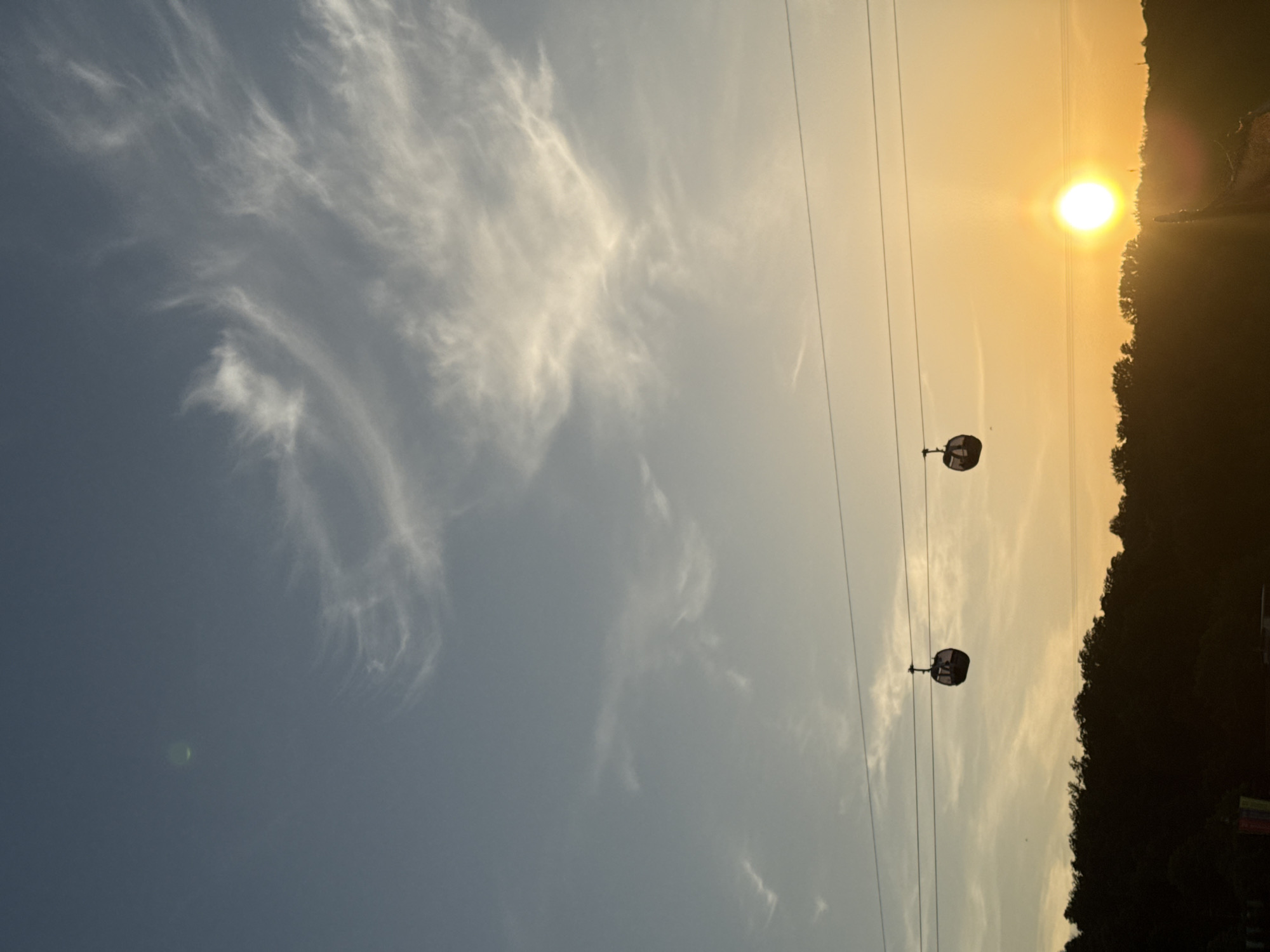 The image depicts a scenic view with a blend of natural and man-made elements, possibly captured during a late afternoon or early evening. In the sky, there are wispy cirrus clouds contrasted with a clear blue segment. The sun is positioned near the horizon, casting a warm, golden light. Two cable cars can be observed, suspended by cables that stretch across the scene. The lower right quadrant of the image shows a dark treeline, likely indicating a wooded area or a park. Based on the presence of cable cars and the general landscape, the location is plausibly Moscow, Russia, specifically in the vicinity of Vorobyovy Gory (Sparrow Hills), where there is a cable car line. There are no people visible in the image, and the scene suggests a quiet, peaceful setting.
