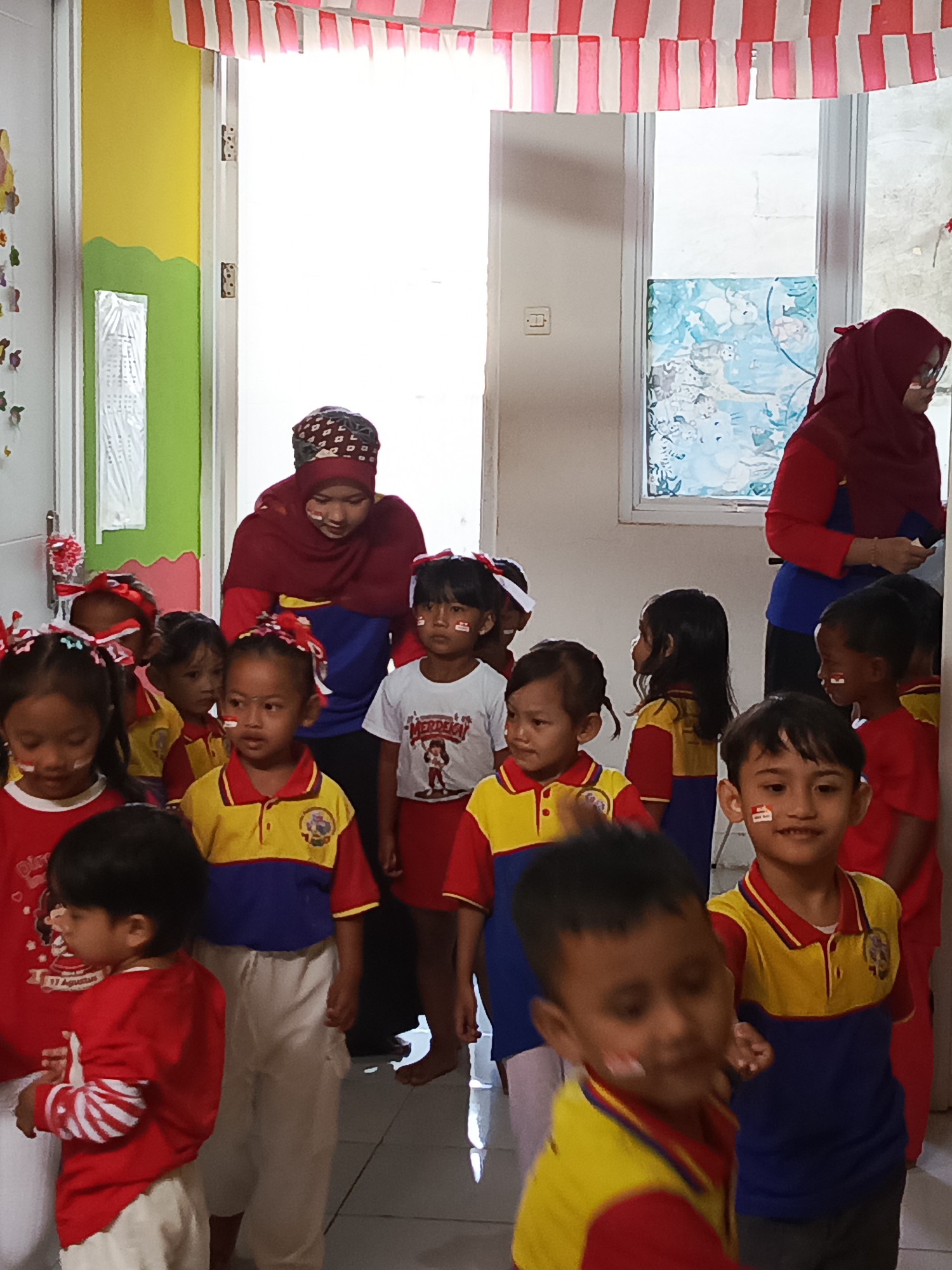 This is a medium shot, eye-level, outdoor, natural-light photograph of a group of young children and two adult women in what appears to be a school hallway or classroom setting in Ciseeng, Indonesia. The children, mostly under the age of ten, are arranged in a cluster, some in the foreground and others in the mid-ground. They are wearing red and white face paint, red and white hair accessories, and some are in school uniforms. One child in the foreground wears a red long-sleeved shirt with white stripes on the sleeves. Another child in the mid-ground wears a red, yellow, and blue collared shirt and white pants. A third child in the mid-ground wears a white t-shirt with an illustration and text, including the word "MERDEKA".
Two adult women are present. One woman in the background wears a dark red headscarf and a blue and red top. She is looking down at the children. The second woman, to the far right, also wears a dark red headscarf and appears to be wearing glasses. She is looking towards the children.
The environment includes a doorway in the background with white and red striped bunting hanging across the top, suggesting a celebration, likely Independence Day given the "MERDEKA" text. The walls of the room are painted in yellow and green. A window with blue and white patterned curtains is visible to the right. The floor appears to be a light-colored tiled surface. The overall atmosphere is festive and focused on the children.