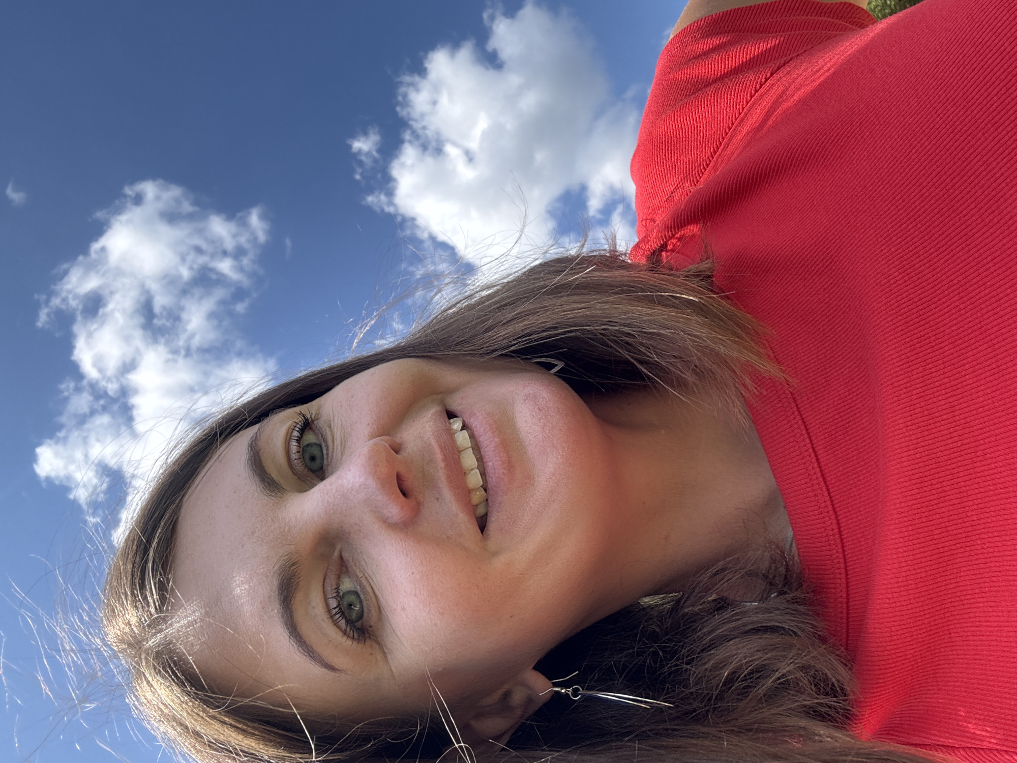 This is a close-up selfie of a woman taken outdoors against a bright blue sky with scattered white clouds. She has brown hair and green eyes, and she is wearing a red top. Her head and shoulders are visible, and she is looking towards the camera with a smile. The image is taken from a low angle, looking up towards the sky. There is no indication of the location being Coteaux du Lizon, France.