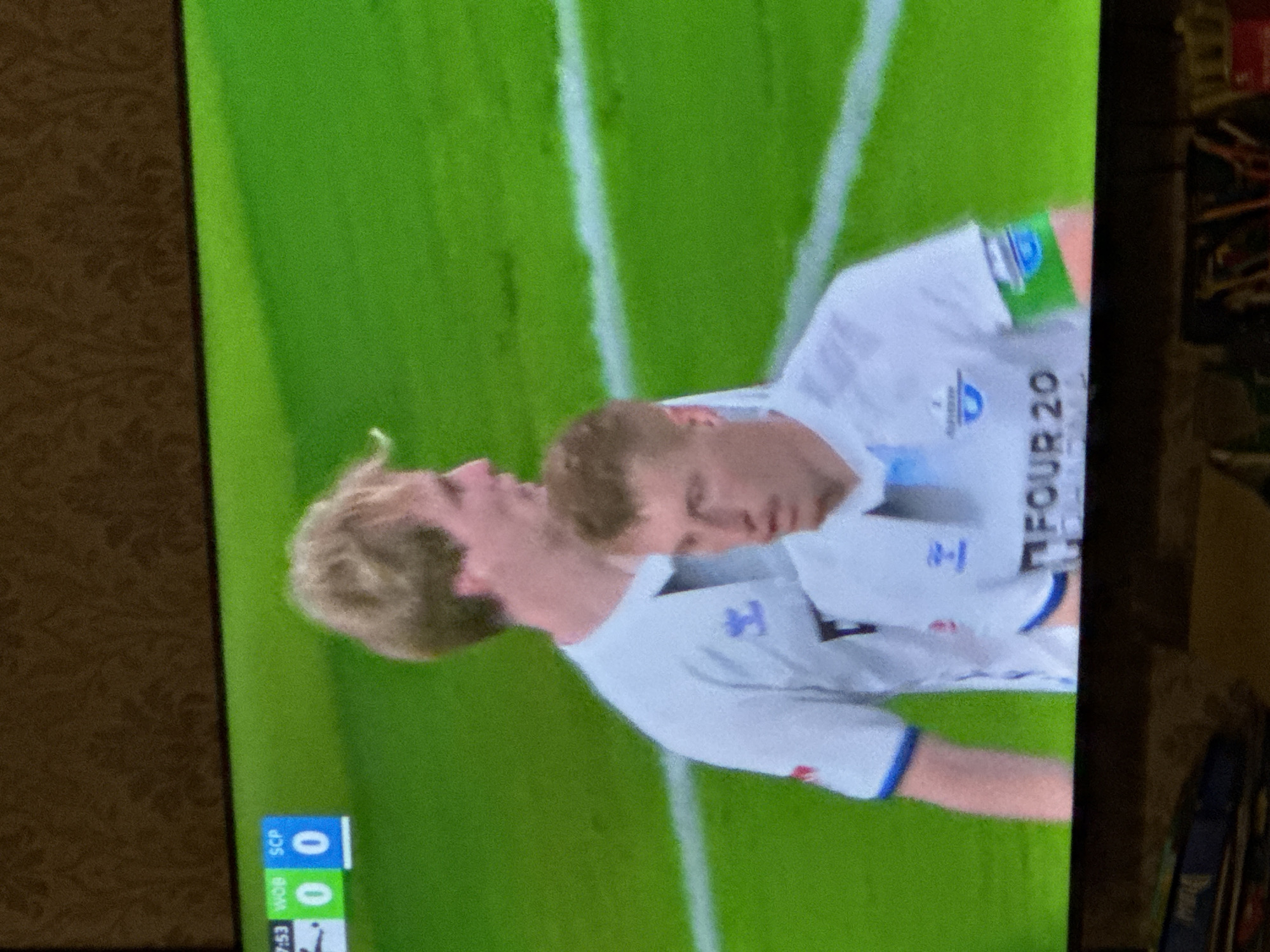 The image shows a television screen displaying a soccer game. Two players in white jerseys with blue trim are visible on a green field with white lines. One jersey has "FOUR 20" printed on it. A scoreboard in the corner reads "WOB 0, SCP 0, 7:53". The location is not identifiable from this image.