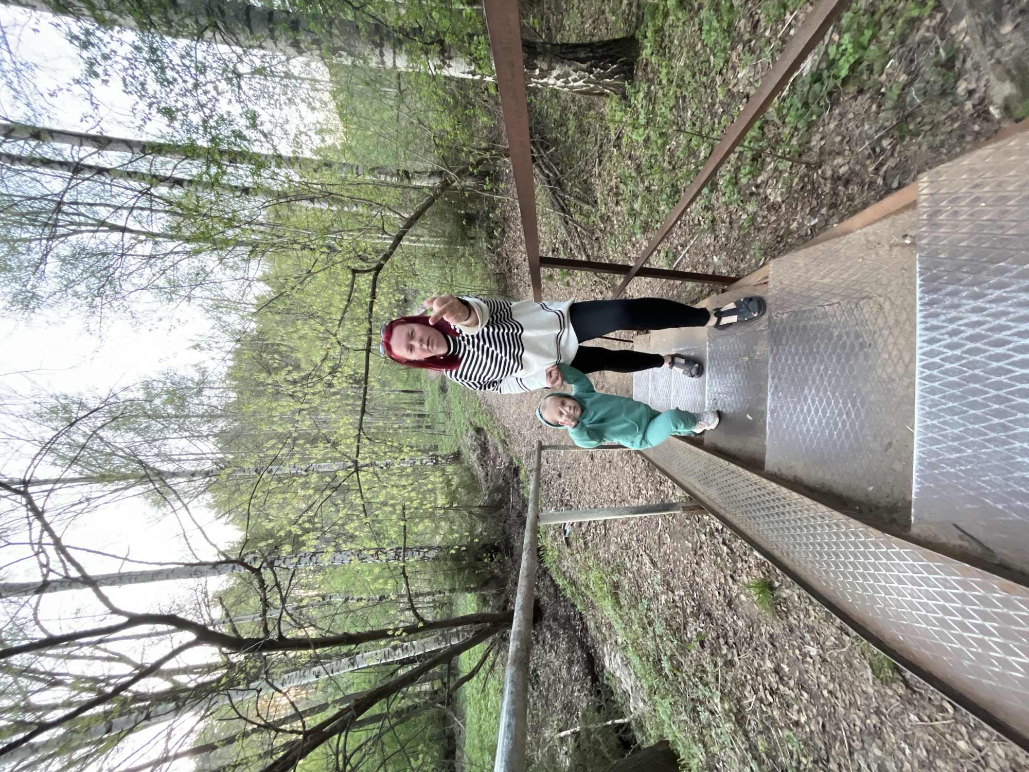 The photograph, taken in what appears to be a wooded area near the city of Chaikovsky, Russia, shows a woman and a child standing on a metal platform with a grated surface. The woman is wearing a striped sweater, dark leggings, and sandals, and she has red hair. She is pointing at the camera. The child is dressed in a green hooded sweatshirt and pants with white shoes. They appear to be holding the woman's hand. The platform is elevated, with forest vegetation visible below. Birch trees surround the platform. The lighting is natural, suggesting a daytime shot.