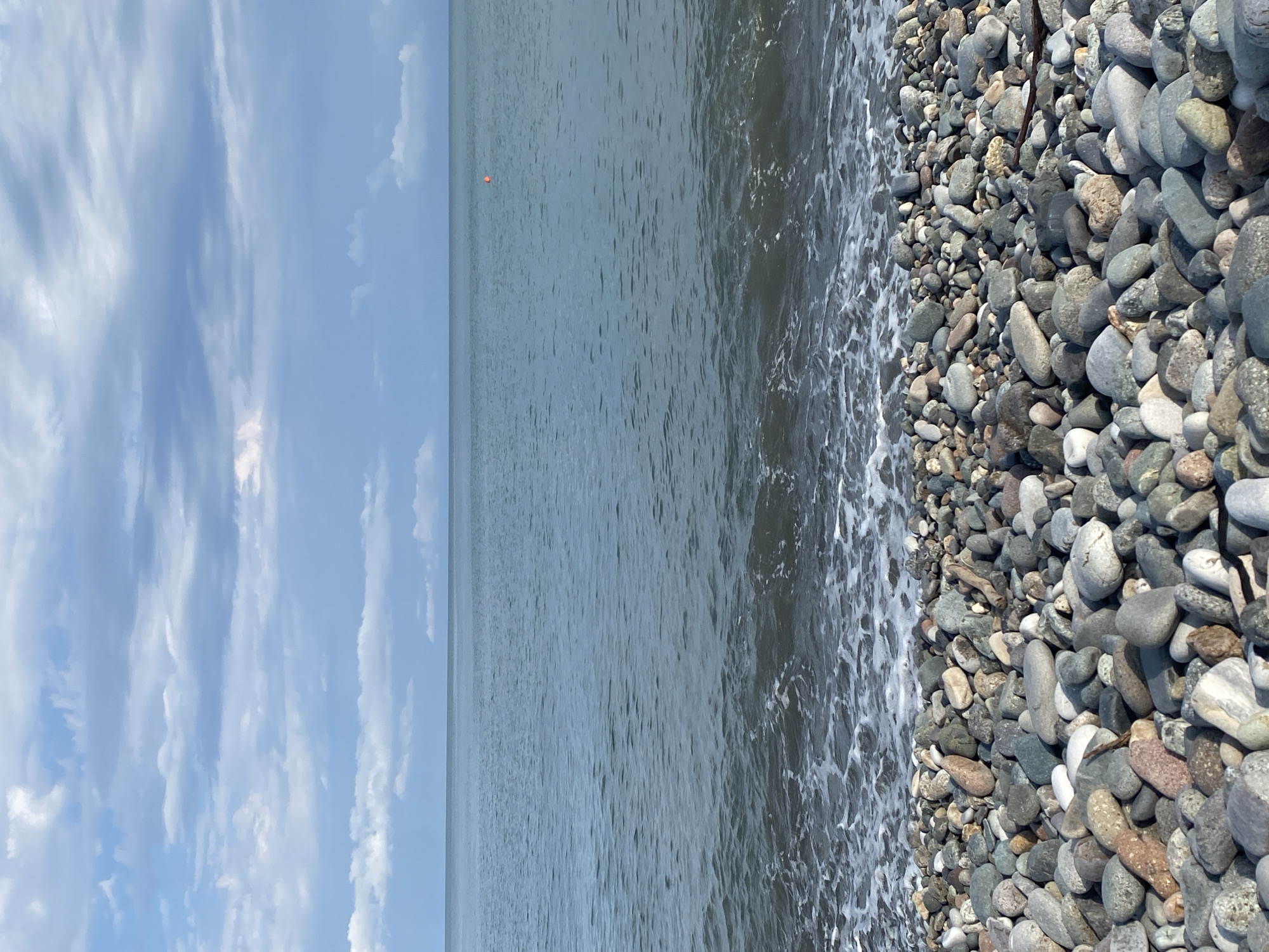 The image depicts a pebbled beach in Batumi, Georgia, with the calm, grey sea stretching towards the horizon under a cloudy sky. Small waves with white foam gently lap at the shore, which is densely covered with smooth, multi-colored pebbles of various sizes. In the distance, a small orange buoy is visible in the water. The sky is filled with layered, wispy clouds in shades of white and grey against a pale blue background. No people or animals are present in the frame.