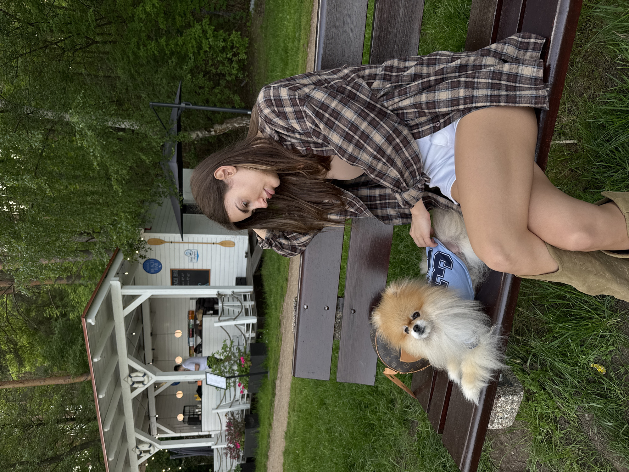 A woman sits on a wooden bench with a small dog dressed in a blue jersey next to her. The woman is wearing a plaid shirt, white shorts, and boots. In the background is a white building with a sign that says "Beach Bar." The surrounding environment features grass and trees. The location is unspecified.
