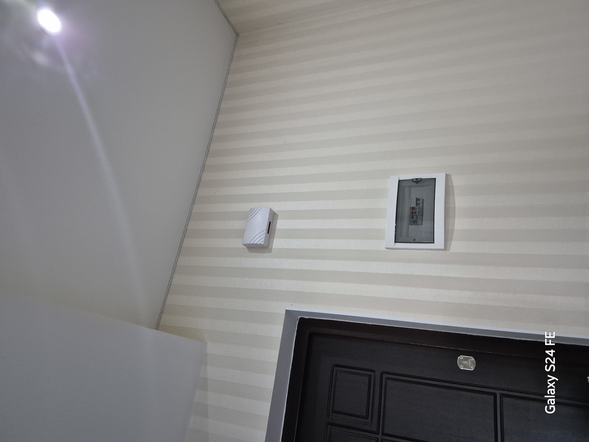 The image shows an interior scene, likely a hallway, featuring a dark-colored door on the left and a wall with vertical stripes of cream and light beige wallpaper. Mounted on the wallpaper are two electrical components: a white rectangular box with a subtle pattern towards the top, and below it, a white framed electrical panel with a transparent cover revealing circuit breakers. The ceiling is white and appears to be a stretch ceiling, with a light fixture visible in the upper right corner casting a bright glare. The overall impression is of a well-lit, clean, and modern interior space. No people or specific identifying markers for Chișinău, Moldova are present.
