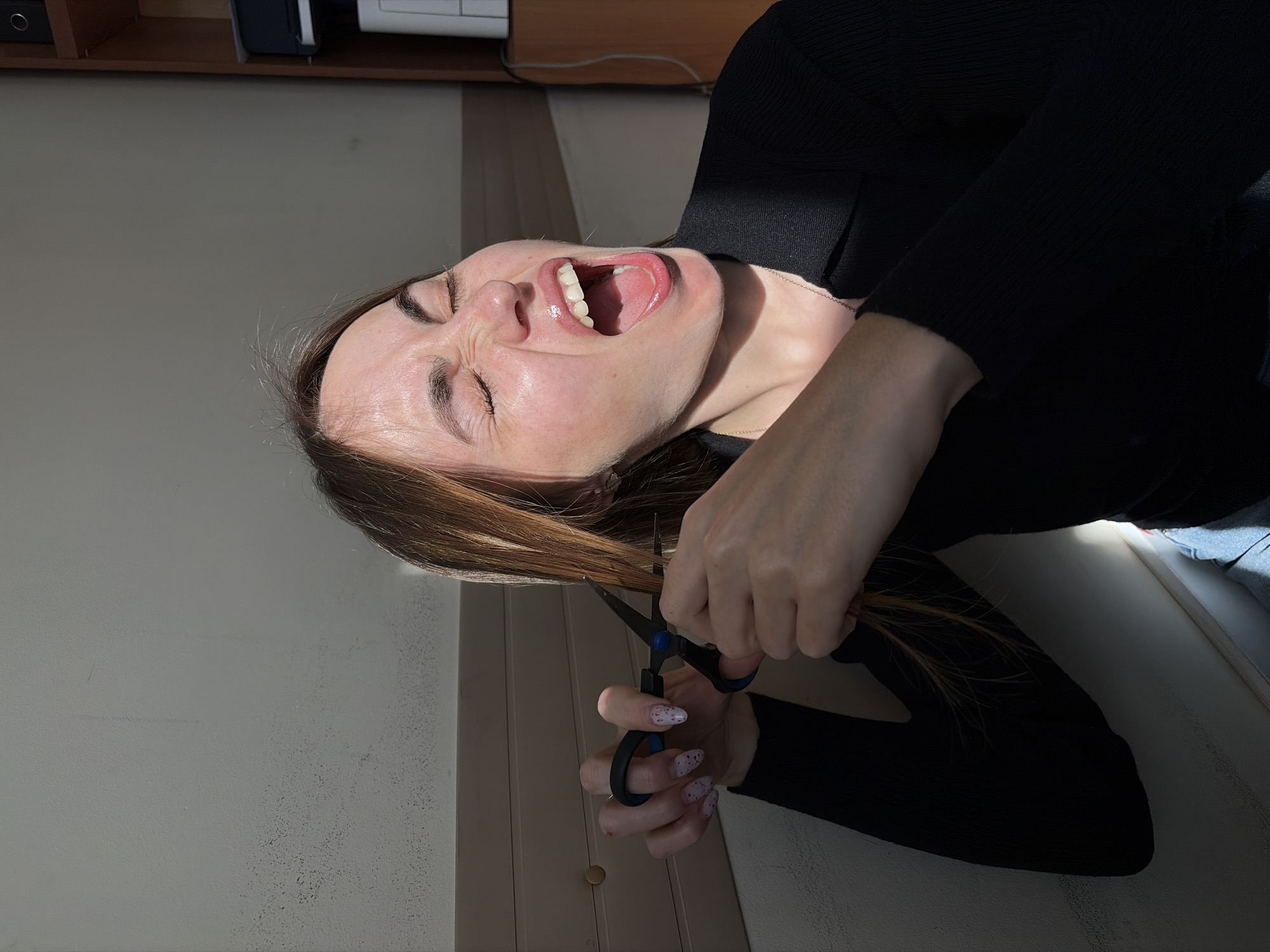 A woman is cutting her hair with scissors in what appears to be an indoor setting. She is wearing a black top and her face is contorted with her mouth open wide and eyes closed, possibly indicating distress or frustration. The location is undetermined; no features present in the image confirm that it is Izhevsk, Russia. A light-colored wall is visible in the background, as well as a cabinet and an electrical cord.