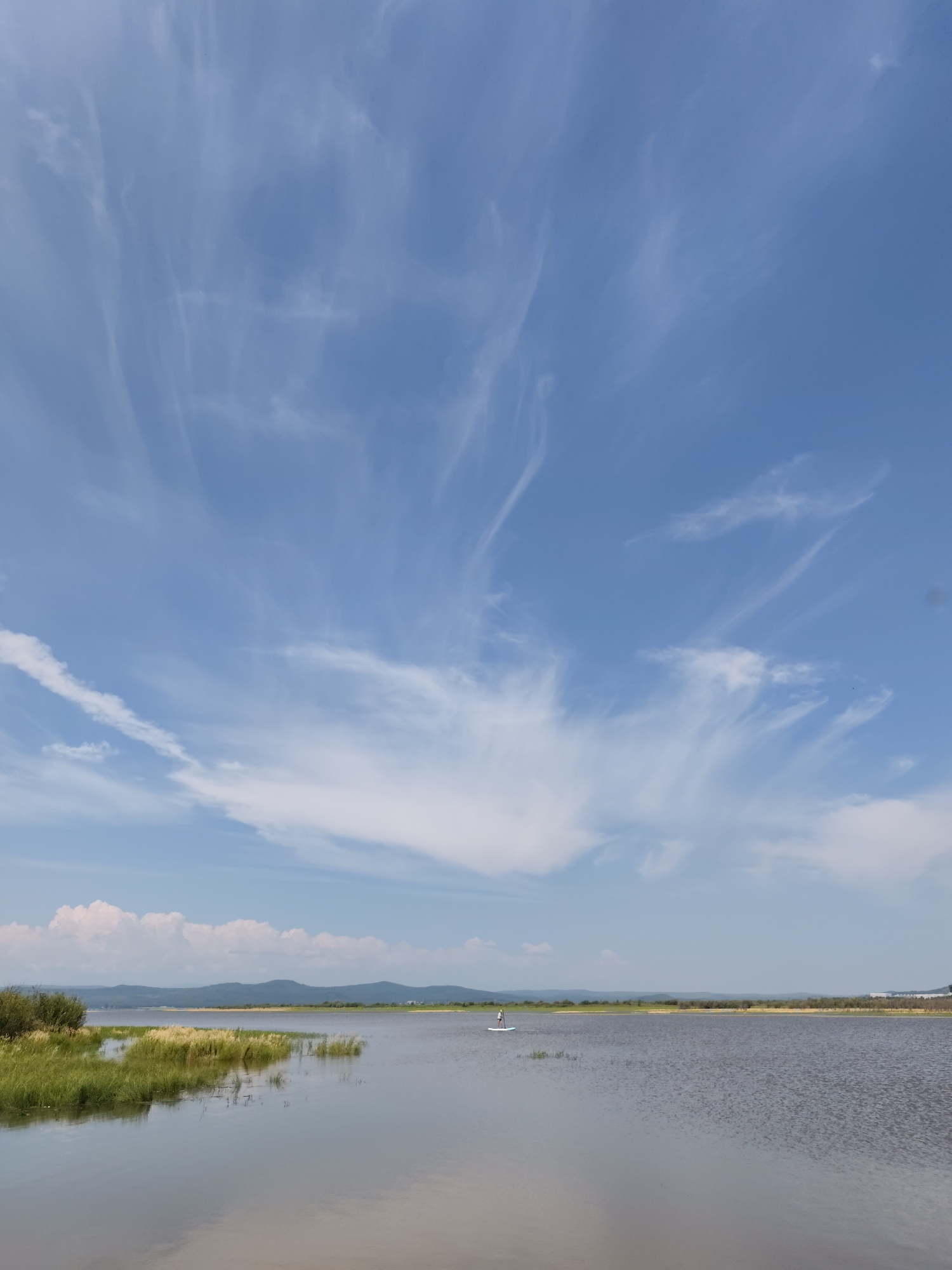 This is a photograph of a wide, calm body of water with a person on a paddleboard in the distance. The water reflects the sky and the surrounding landscape. The foreground shows a marshy area with green and yellow grasses. In the background, there are low, rolling hills or mountains covered in vegetation. The sky is a clear blue with scattered, wispy white clouds. The overall impression is one of a serene natural landscape. Based on the available information about Komsomolsk-on-Amur, Russia, this scene is likely a view of the Amur River or one of its tributaries or lakes in the vicinity of the city, possibly during the warmer months. The light suggests it is daytime, likely late morning or afternoon.