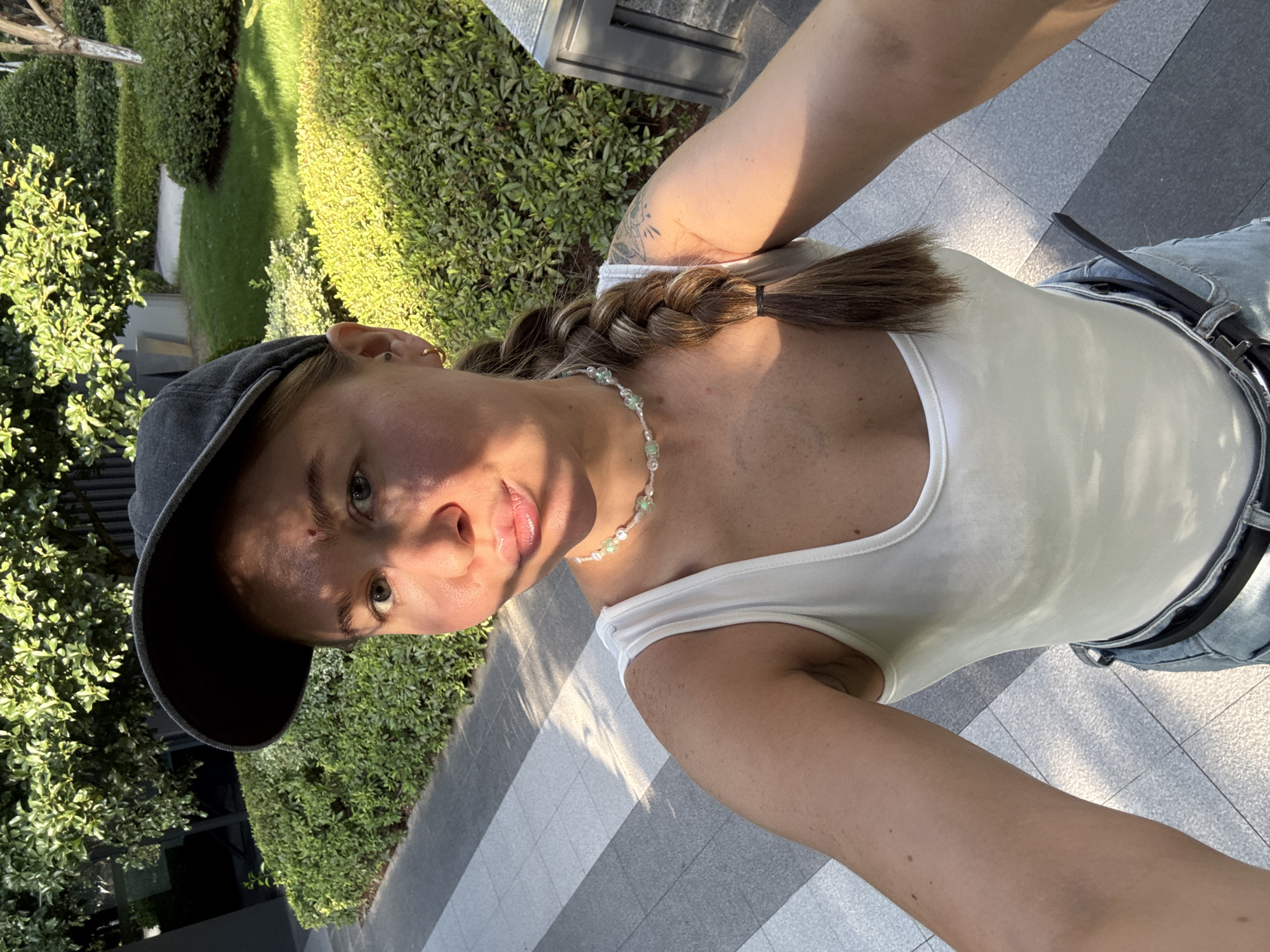 A young woman wearing a white tank top, a black baseball cap, and blue jeans poses for a selfie outdoors. She has a braid in her hair and is wearing a beaded necklace. The background features manicured green bushes and a tiled walkway. The scene appears to be a park or courtyard area in Yiwu, China, with buildings and trees visible in the distance. The lighting suggests it is daytime, with the sun casting shadows on the ground.