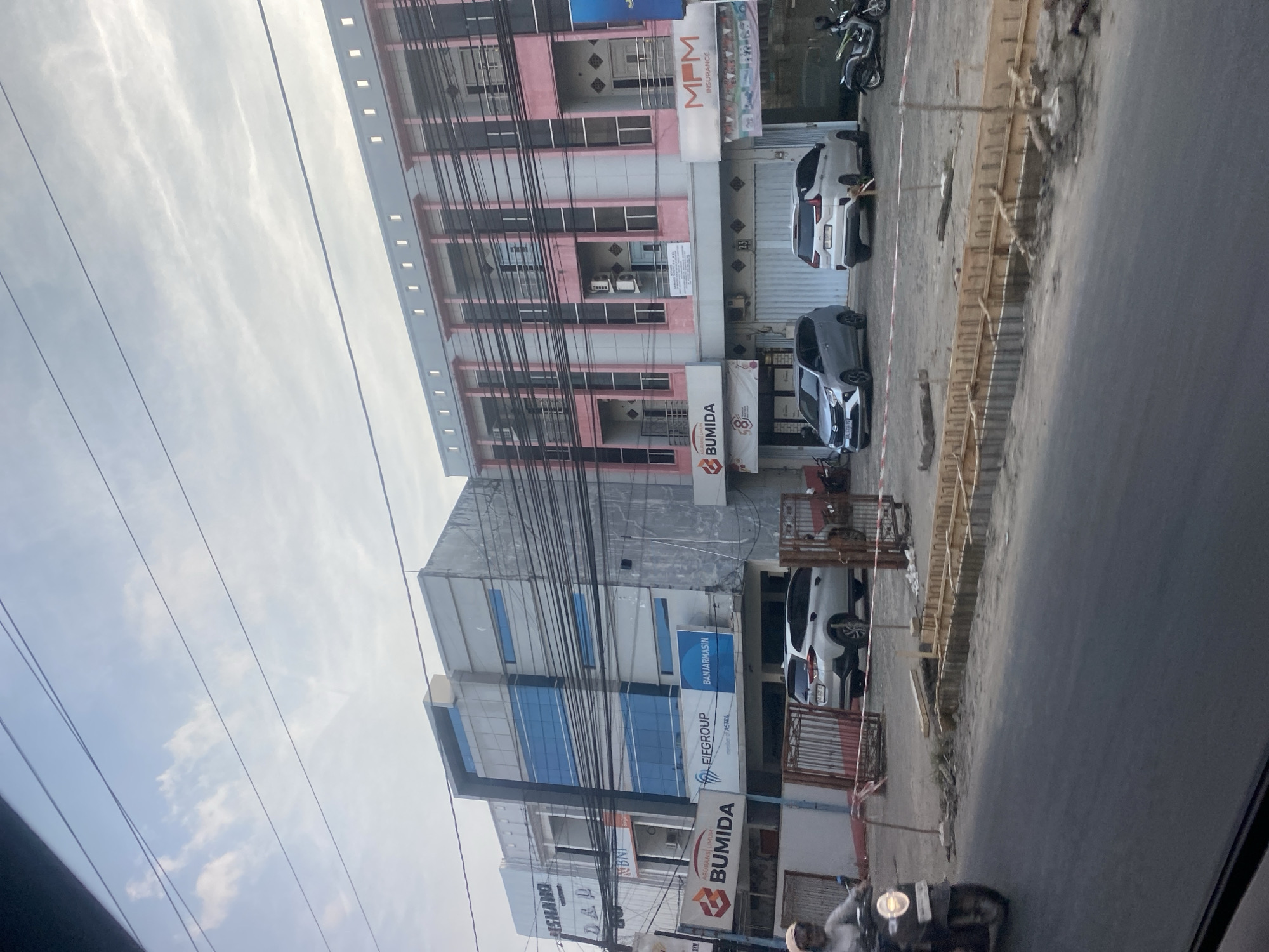 This image captures a street view in Banjarmasin, Indonesia, featuring a row of multi-story buildings. The buildings exhibit modern architecture with elements like glass facades, balconies, and visible air conditioning units. Signs on the buildings indicate various businesses, including "BUMIDA," "EIGGROUP," "BNI," and "MPM INSURANCE." Several vehicles are parked in front of the buildings, including a silver SUV, a silver sedan, and a motorcycle. A person is riding a motorcycle on the street in the foreground. Construction materials, including wooden forms and rebar, are visible along the roadside, suggesting ongoing infrastructure work. Overhead power lines crisscross the sky, and the sky itself is partly cloudy. The overall scene depicts a commercial area with ongoing development.