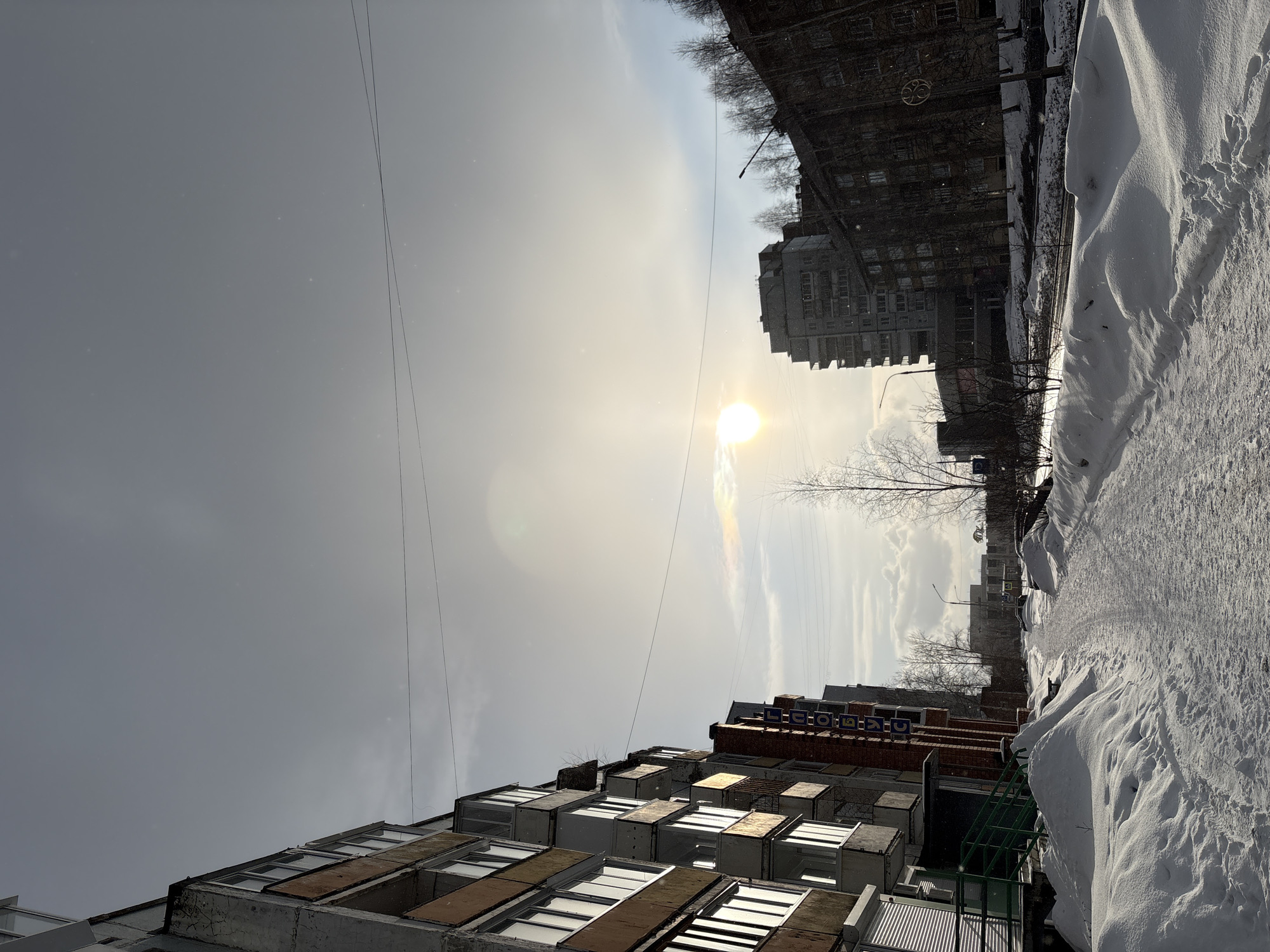 The image captures a winter scene in what appears to be a residential area of Ust-Ilimsk, Russia. The viewpoint is from the ground level, looking up at buildings under a bright, hazy sun. Snow is heavily present along the side of what looks like a sidewalk or road.
The buildings are typical Soviet-era apartment blocks, with multiple stories and uniform window designs. Some windows are boarded up. One of the buildings features a sign in Russian, which appears to be the name of a store or organization. Utility wires stretch across the sky. There are trees between the buildings, most of which are bare due to the winter season. The sky is overcast, but the sun shines through the clouds, creating a glare in the image. No people are visible in the frame.