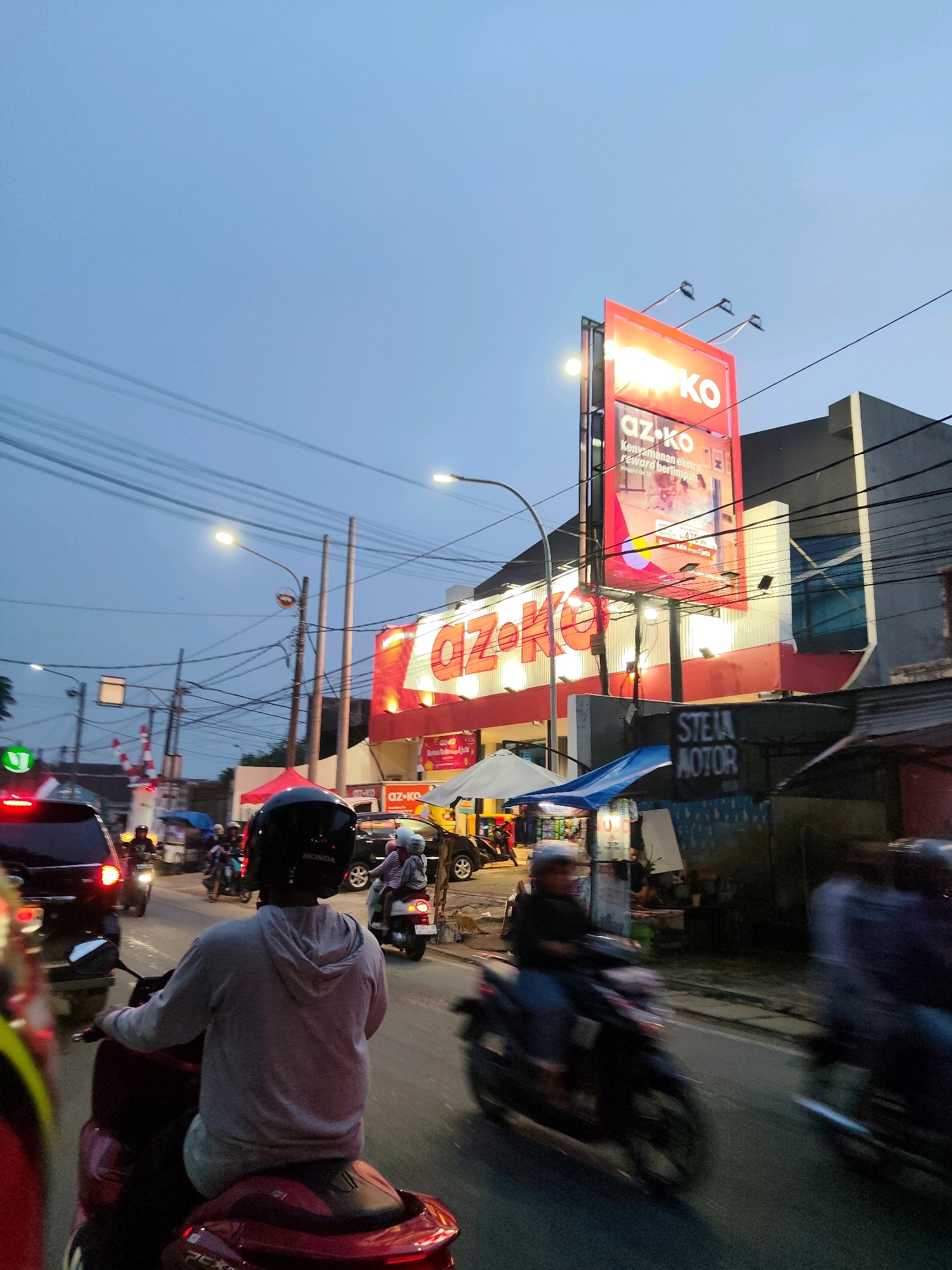 The image captures a street scene at dusk in Kaduagung Timur, Indonesia, with a prominent red and white sign for "az-ko" illuminated against a dusky blue sky. The "az-ko" sign is large, rectangular, and positioned above a building with similar red and white branding. Below the main sign, a smaller section of the "az-ko" branding is visible, with text that appears to be promotional. The street is busy with vehicular traffic, primarily motorcycles. Several individuals are riding motorcycles, some with passengers. One person in the foreground, facing away from the camera, is wearing a grey hoodie and a black helmet, riding a red scooter. To their right, another person on a motorcycle is also moving forward. Further down the street, more vehicles and people are visible, some blurred due to motion. A railroad crossing gate is visible on the left side of the image, with red and white striped barriers. Utility poles and wires are strung across the sky, characteristic of an urban or semi-urban environment. The overall impression is a bustling street scene during the transition from daylight to evening.