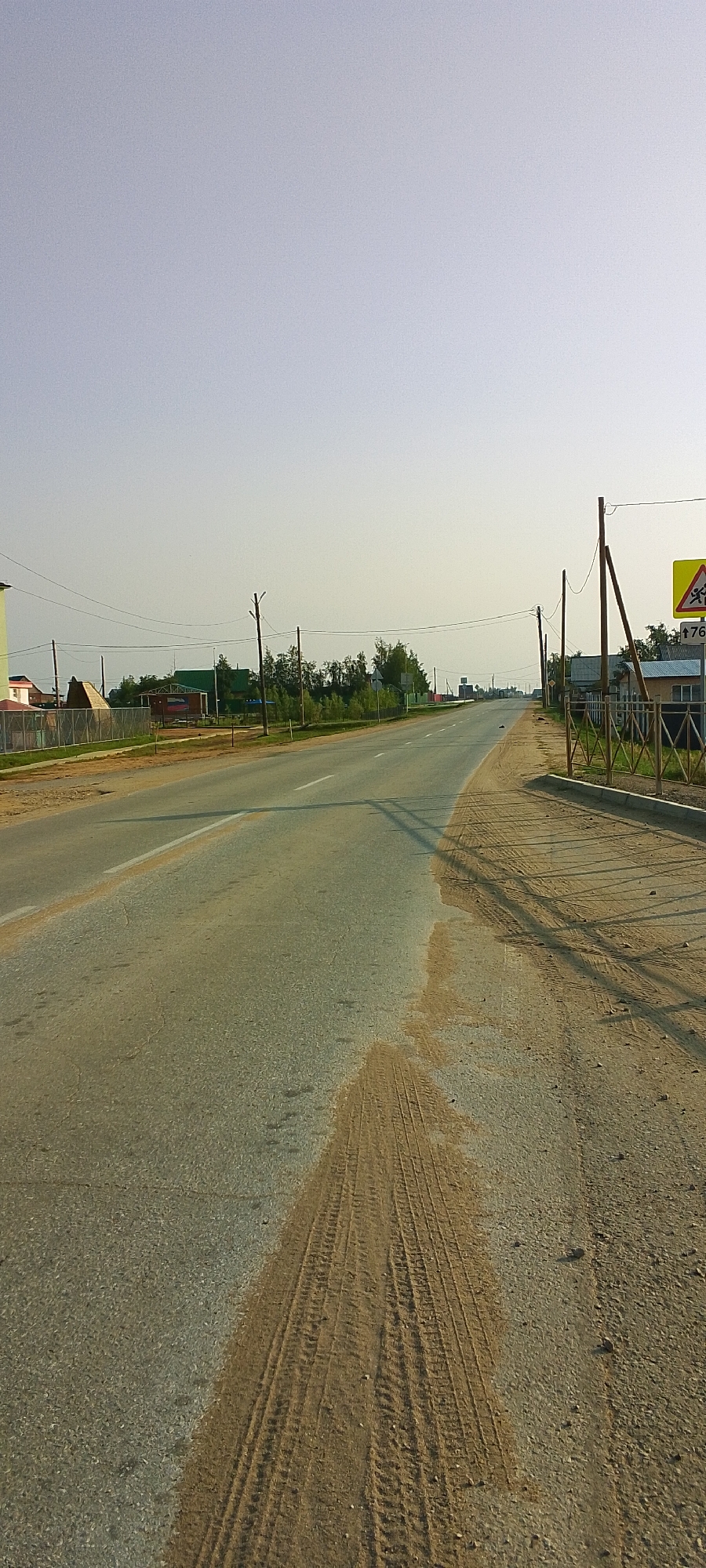 The image shows a straight, paved road running through a rural or suburban area, likely in Tulagino, Russia. The road is bordered by a dirt shoulder on the right, which has tire tracks from a vehicle. Along the left side of the road, there are several houses with various architectural styles and fence lines. Trees and utility poles are visible on both sides of the road. In the distance, the road continues to curve slightly. The sky is clear and light blue. A yellow triangular road sign with a symbol of a person walking and the number "76" is visible on the right side of the road. The overall impression is a view of a typical day in a Russian settlement.