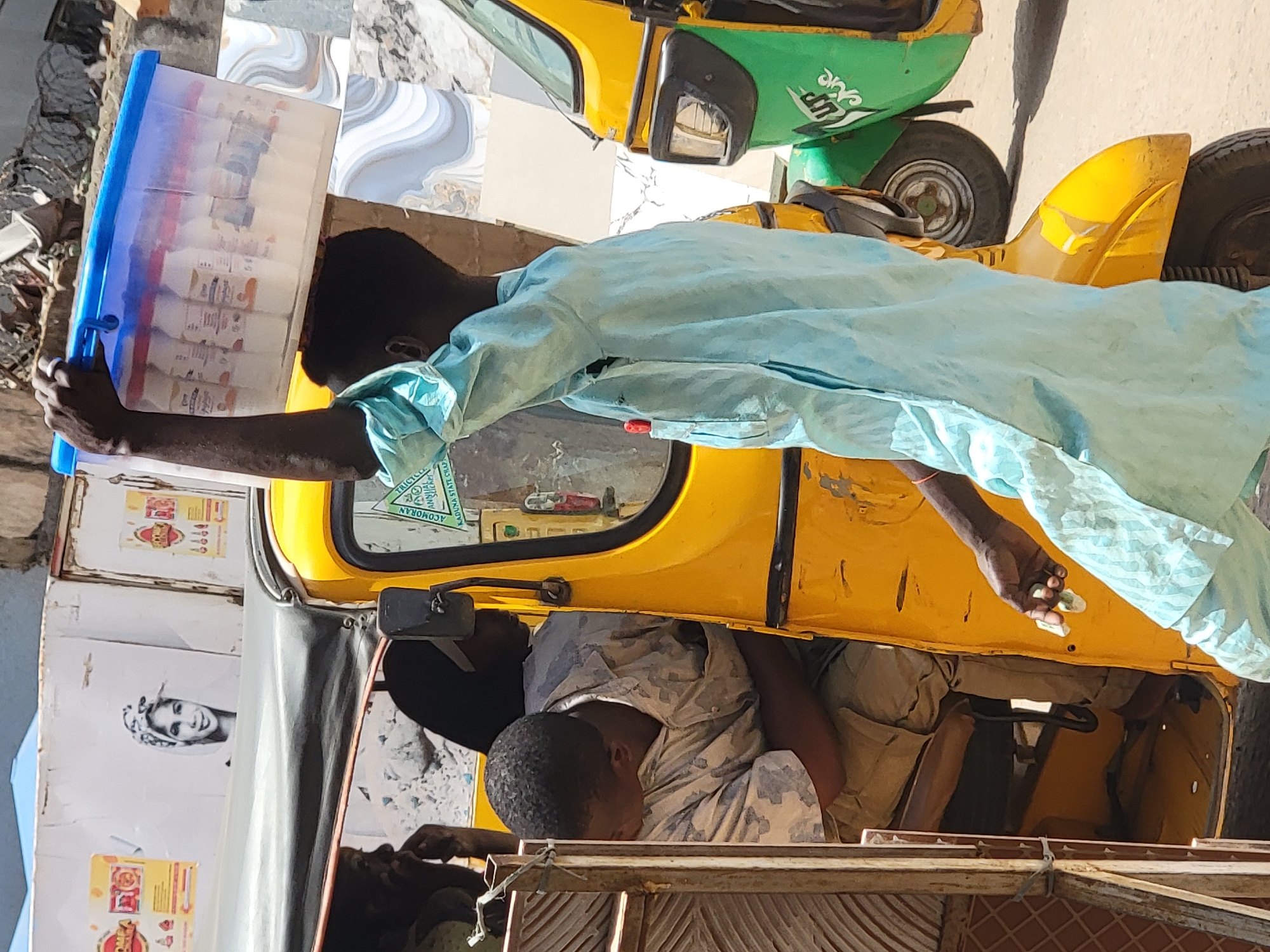 The image, rotated 90 degrees clockwise, depicts a bustling street scene in Maiburiji, Nigeria, under bright daylight. In the foreground, a person with dark skin, wearing a light blue long-sleeved garment, holds a large, clear rectangular plastic container with a blue lid and handle. This container is filled with numerous smaller, individually packaged items, likely snacks or beverages offered for sale by the vendor. Their left hand appears to be holding a small, light-colored object, possibly money or an item being exchanged.
A prominent yellow tricycle, often referred to as a Keke Napep, occupies the central part of the frame. Inside, another person with dark skin is seen resting or sleeping, dressed in a light-colored, possibly patterned top and dark trousers. The interior shows parts of the vehicle's controls. To the left of the yellow tricycle, stacked cardboard boxes are visible, one prominently displaying a black and white illustration of a woman's face.
In the background, a portion of a green and yellow tricycle can be seen, including its wheel and headlight, situated behind the yellow one. The ground surface in various parts of the image features an assortment of patterned tiles.
Visible text includes "TRICYCLE" and "ACOMORAN" on a sticker inside the yellow tricycle, and "UP" along with some stylized script in white on the green tricycle. This scene captures a typical daily street vending activity, characteristic of urban life in the region.