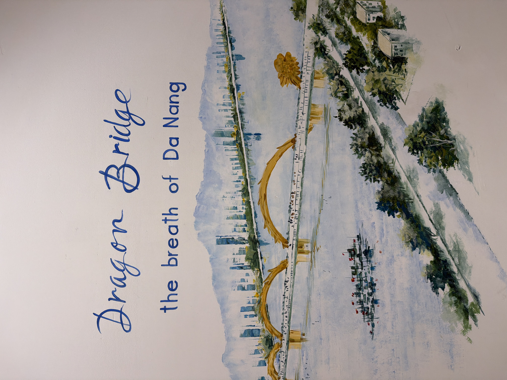 This image is a painting depicting the Dragon Bridge in Da Nang, Vietnam, spanning a river. The text above the painting reads "Dragon Bridge the breath of Da Nang." The bridge is characterized by its golden arches, resembling a dragon. The background features a cityscape with tall buildings and a mountain range. The foreground shows a body of water with several boats, likely fishing vessels, adorned with red flags. The riverbanks are lined with lush green trees and a few small buildings, suggesting a coastal or riverside urban environment. The overall style is impressionistic, with soft brushstrokes and a muted color palette dominated by blues, greens, and yellows.