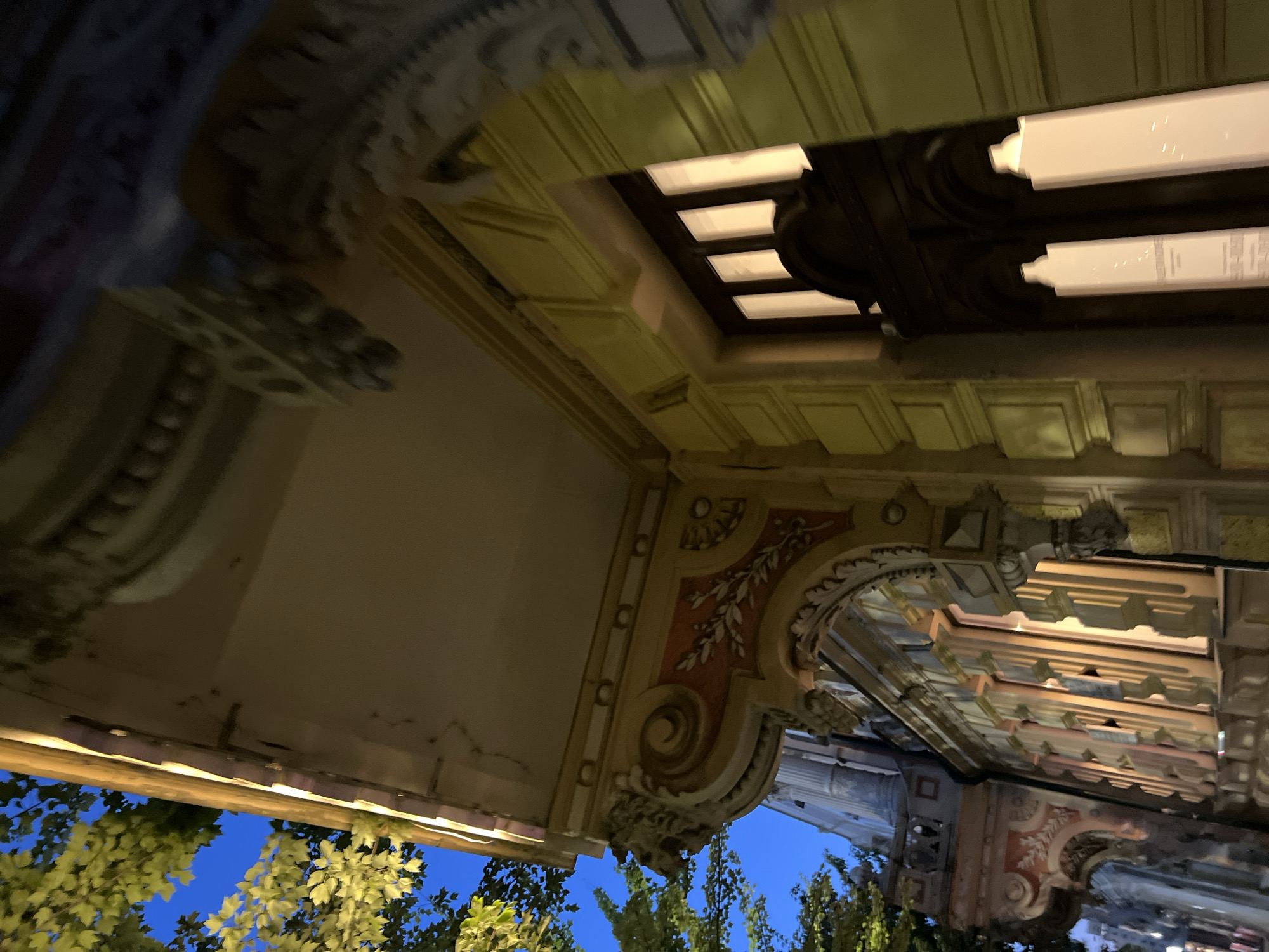The image shows a low-angle shot of the exterior of a building at night in Krasnodar, Russia. The building features ornate architectural details, including sculpted cornices, decorative moldings, and intricate carvings around the windows and doorways. The facade is painted in warm, muted tones of yellow and cream, with accents of red and gold. The windows are lit from within, casting a warm glow onto the building's exterior. Lush green foliage from trees is visible in the upper left corner against a deep blue night sky. The overall impression is one of grandeur and historical significance.