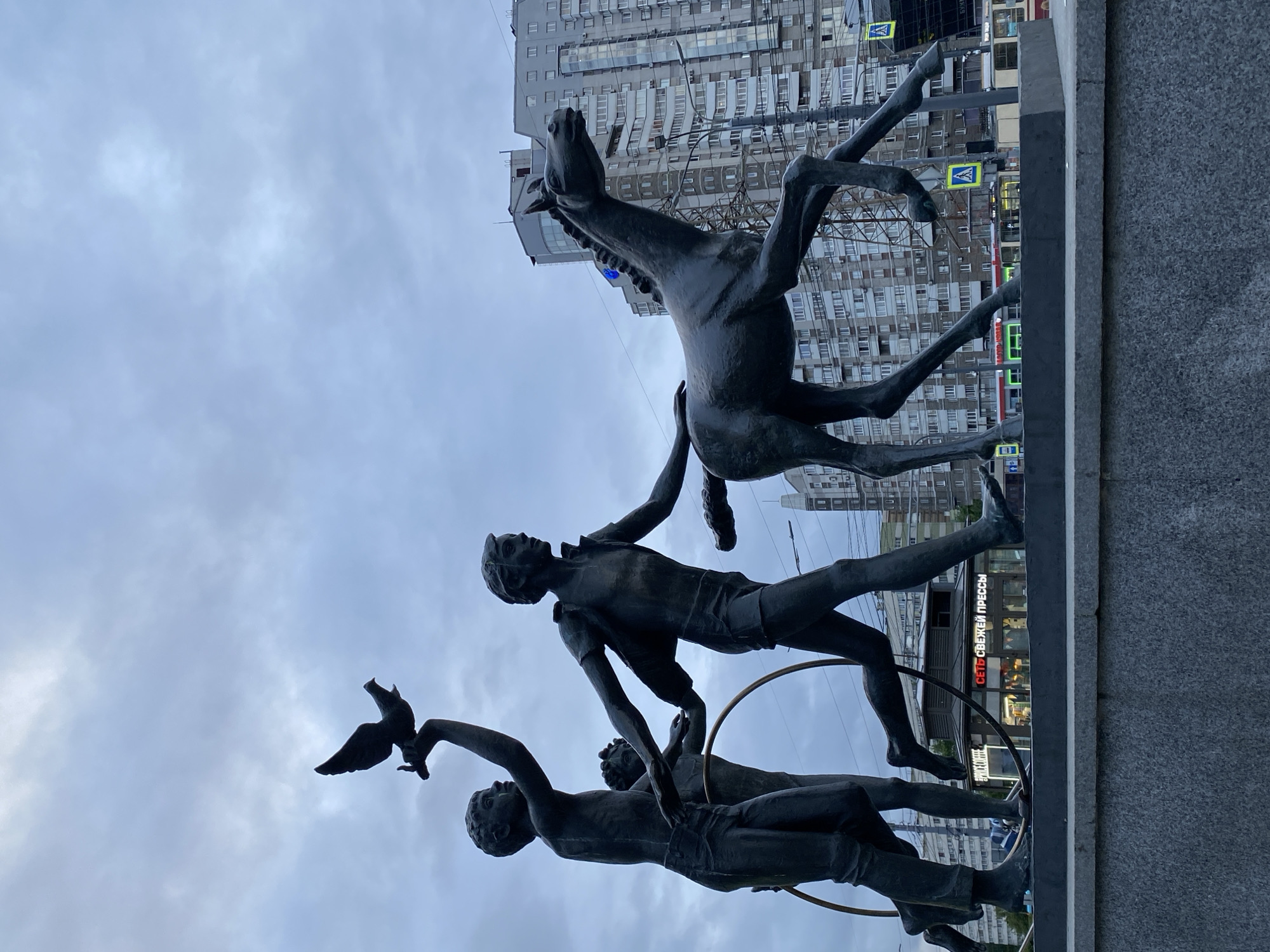 The image shows the "Children of the 21st Century" monument in Saint Petersburg, Russia. The bronze sculpture features three children and a horse. One child is holding a dove, and another is in mid-air, seemingly jumping through a hoop held by a third child. The horse is also airborne. The backdrop includes a modern apartment building and street signs. The sky is cloudy. The monument is placed on a stone base.