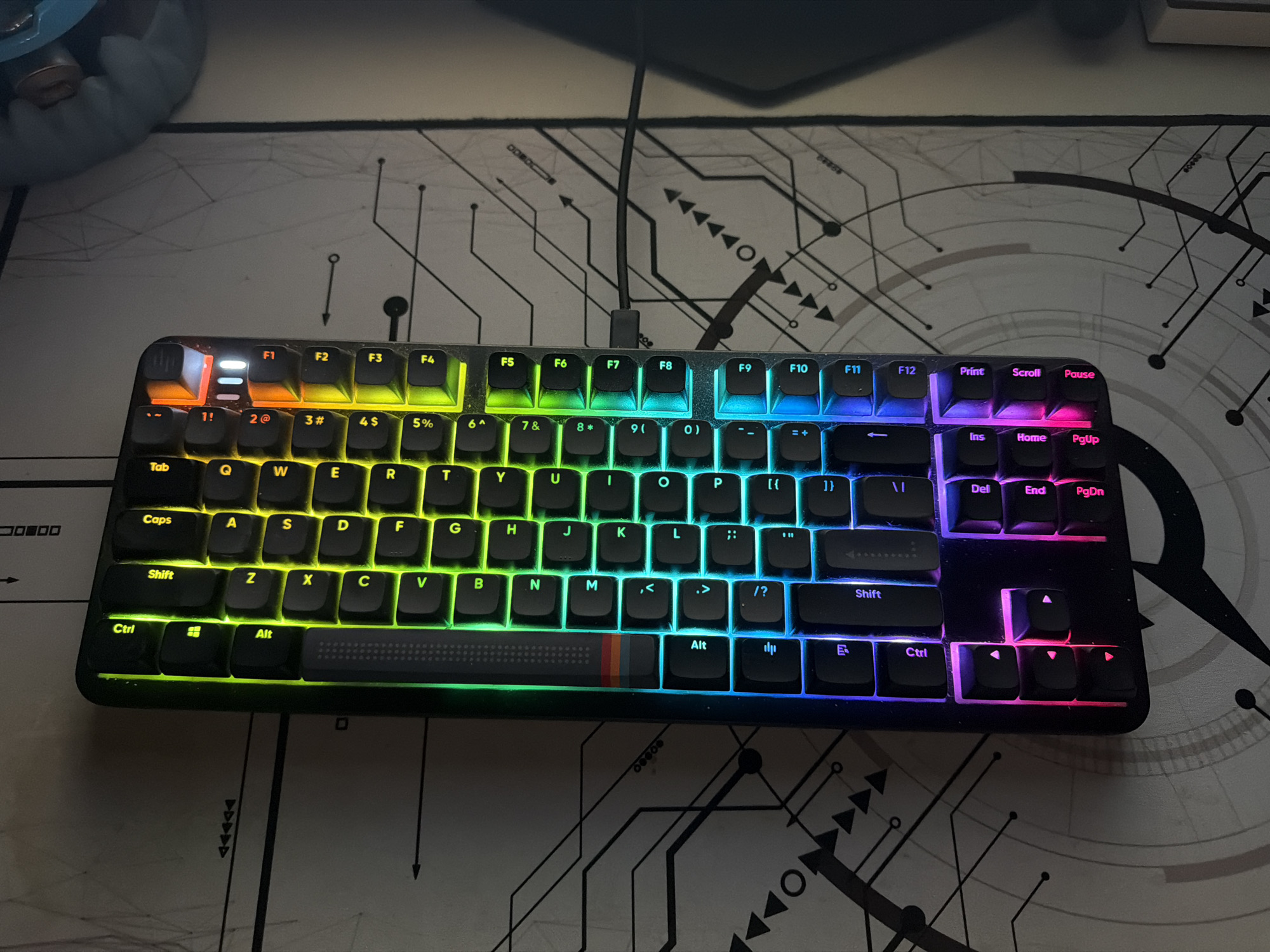 The image shows a mechanical keyboard with rainbow-colored LED backlighting. The keyboard is laid on top of a mousepad with a white and gray circuit board design. The keyboard is a tenkeyless (TKL) layout, with a textured spacebar. The lighting scheme transitions across the keys from red to purple. The function row is marked with F1 through F12. The keys are black with white legends. A black cable is attached to the top of the keyboard. A blue item is partially visible at the upper left corner of the image. The scene is indoors. The location is not provided and is not discernible from the image.