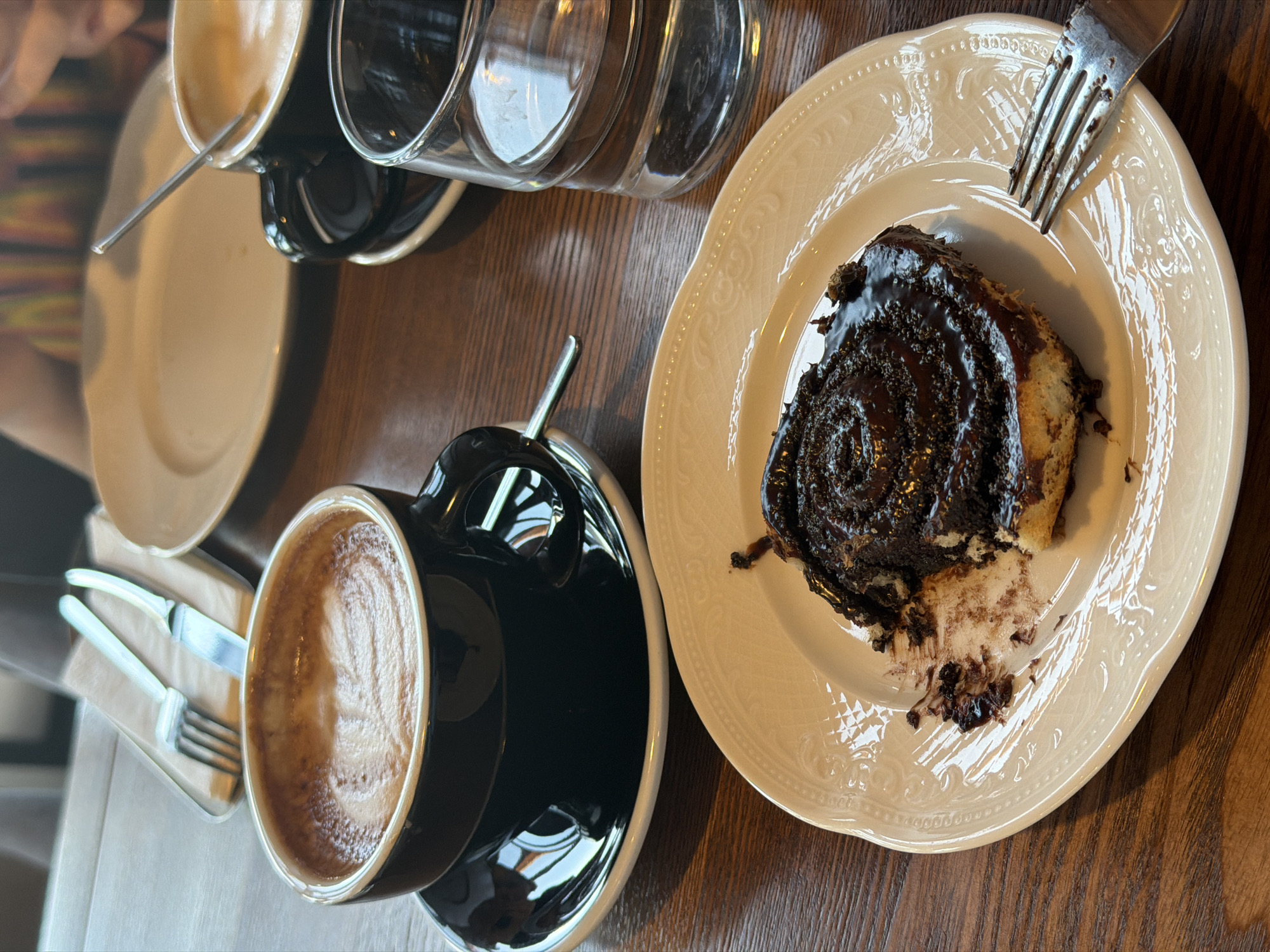 A top-down shot captures a table setting, likely in a cafe or restaurant in Perm, Russia. A black cup filled with latte sits on a saucer. Adjacent to it is a light beige plate with a dark chocolate-covered pastry. A fork rests on the pastry plate. In the background are stacked plates and empty drinking glasses. Cutlery wrapped in a napkin are visible to the left. The table is made of dark wood.