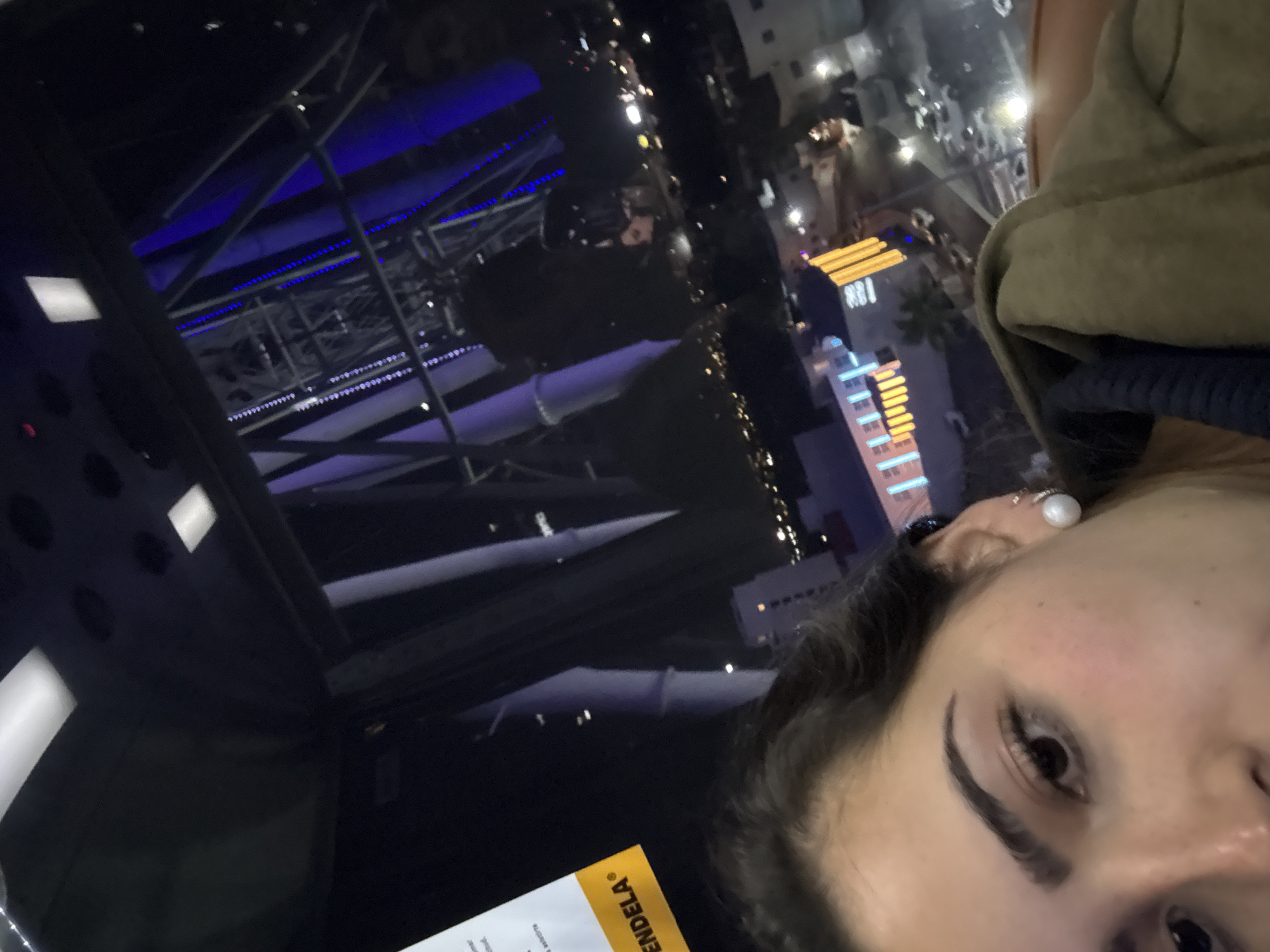 The image shows a person with dark hair and an earring in the upper right, partially framed by an olive green jacket. A menu with the partial word "ENDELA" is visible in the lower left. The background is a blurred cityscape at night, with a dark table reflecting the urban environment. There are visible building structures, including some with blue and orange lighting. The scene appears to be taken from a high vantage point, possibly within a building overlooking the city. The location is possibly in Puebla, Mexico, based on the menu with 'ENDELA'. The interior structure has visible blue lighting.