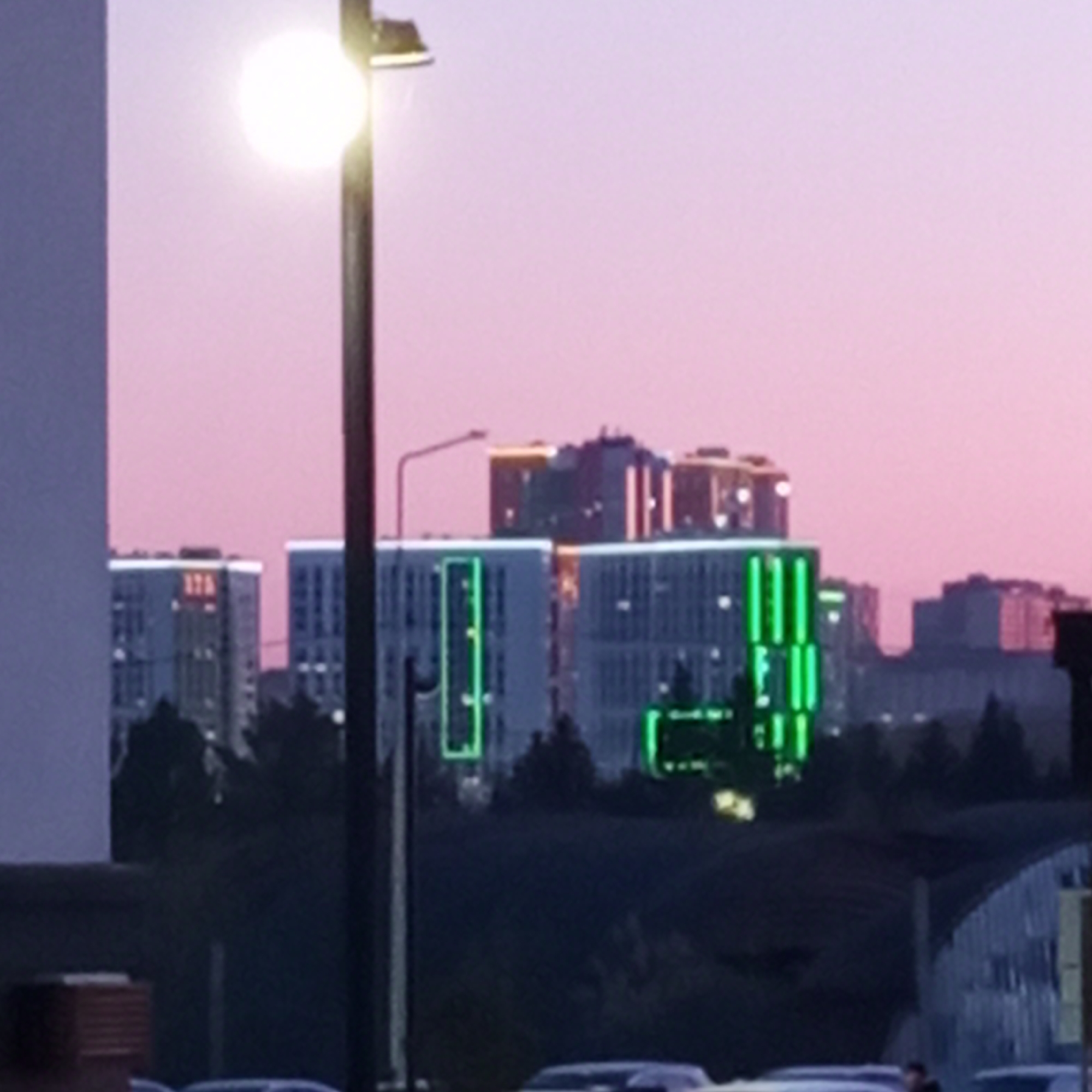 The image presents an urban landscape at twilight, likely either sunrise or sunset, characterized by a soft, gradient sky transitioning from pink at the horizon to light purple higher up. In the foreground, a brightly illuminated street lamp with a circular bulb dominates the left side, its light creating a strong glow.
In the mid-ground, several modern multi-story buildings are visible. Many of these structures feature distinct architectural lighting; some display horizontal bands of warm yellow or orange light near their rooftops, while others, particularly in the central view, are adorned with prominent vertical strips of bright green illumination along their facades. On one building to the left, an illuminated red sign clearly displays the text "17A".
Dark silhouettes of trees and foliage form a natural barrier between the ground level and the buildings. At the very bottom of the frame, blurred shapes suggest parked vehicles, and a partially visible figure of a person can be discerned to the right. The overall scene conveys a serene and contemporary urban atmosphere as artificial lights begin to punctuate the fading natural light. No specific activity is prominently featured.