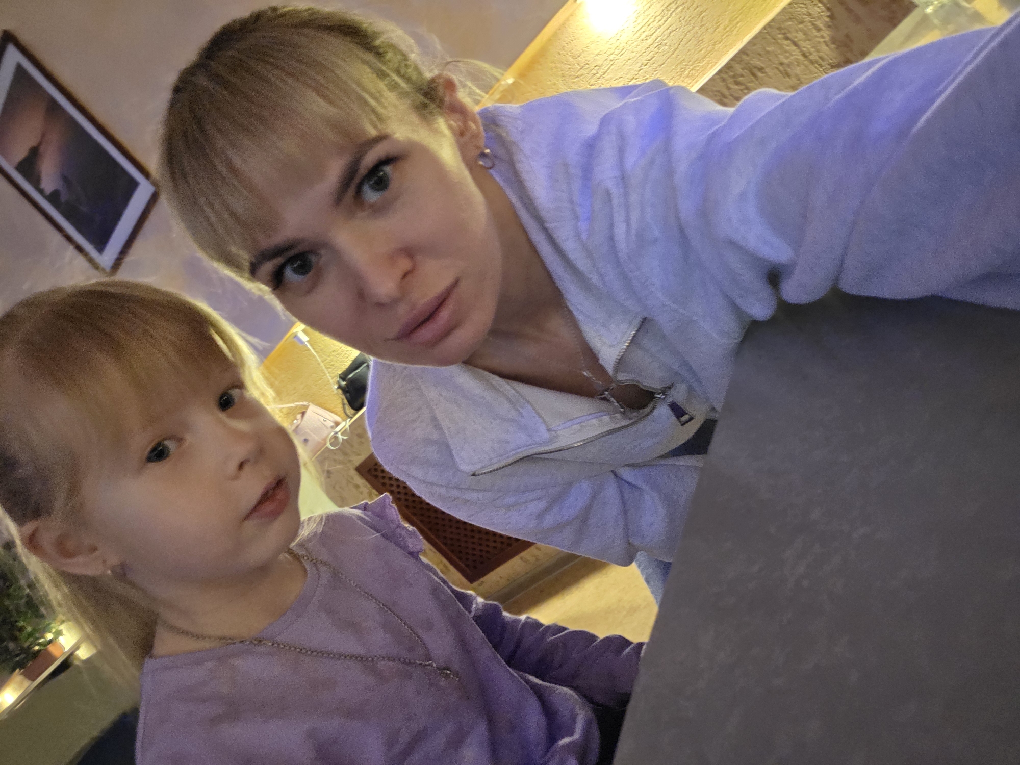 A woman with blonde hair and a young girl, also blonde, are visible in the frame. The woman has her hair pulled back, with bangs, and is wearing a light-colored zip-up sweatshirt. The young girl is wearing a lilac-colored long-sleeved shirt with a small necklace. A framed picture hangs on the wall in the background. The location is Chaikovsky, Russia.