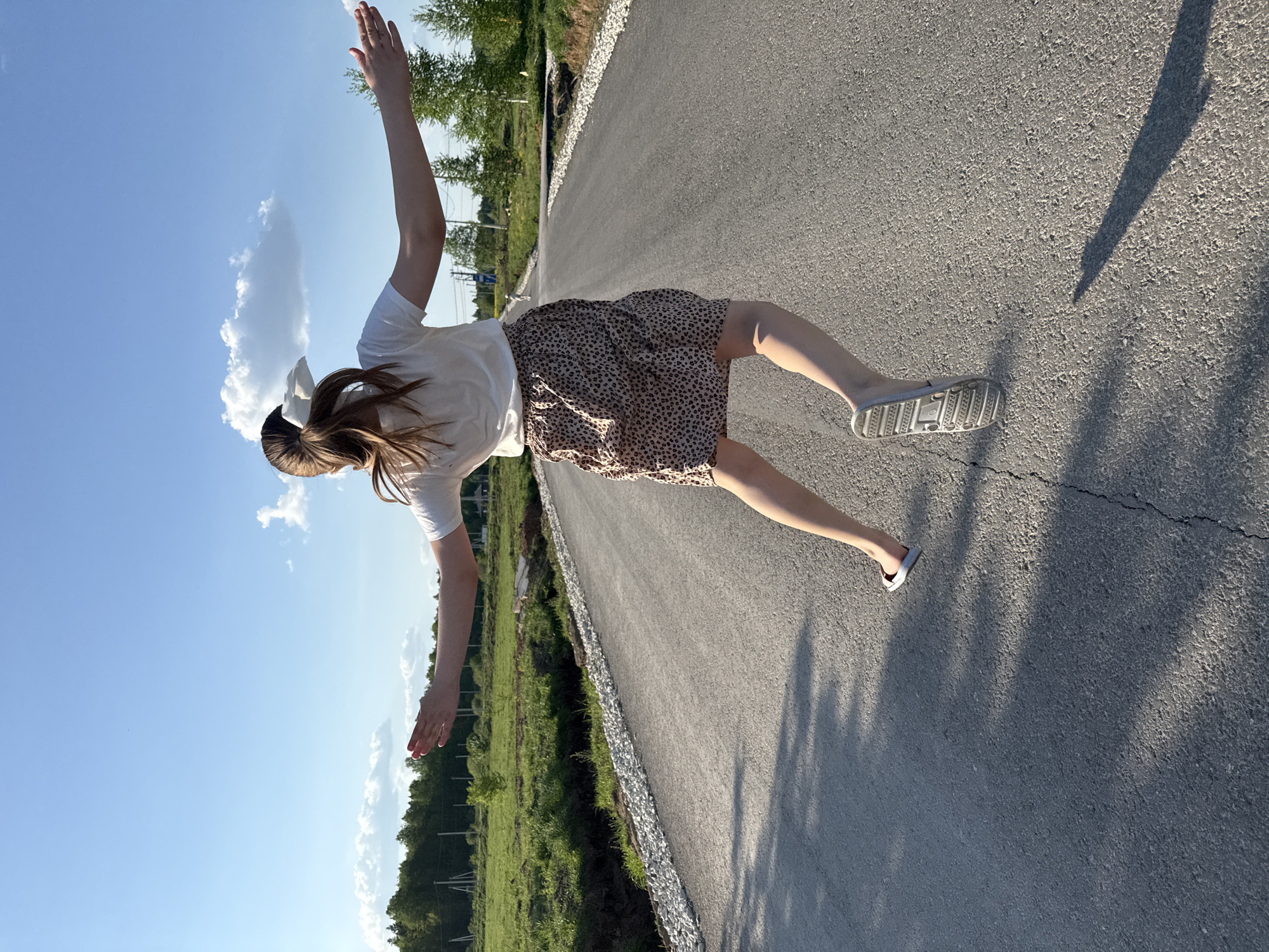 A female subject with long brown hair, wearing a white t-shirt, a brown and black spotted skirt, and gray sandals, stands on a paved road with her arms outstretched. She is oriented with her head toward the sky, which is visible above, and her feet toward the road. The road is bordered by green vegetation on one side and appears to be newly constructed with white gravel along the edge. The photograph gives the impression of levitation, but it is due to the camera's angle. The location is potentially in Izhevsk, Russia, but no specific landmarks confirm this.