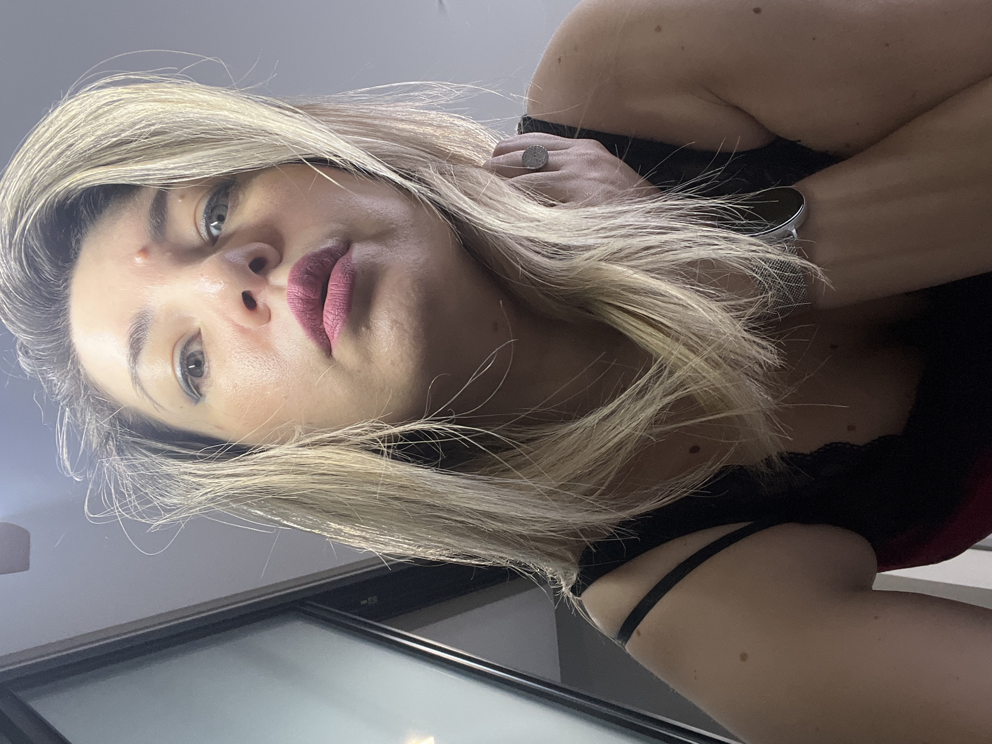 The media file is a low-angle, close-up shot of a woman's face and upper body, taken indoors. The woman has blonde hair, styled with some strands falling across her face, and is wearing makeup, including lipstick and eye makeup. She is also wearing a black garment with lace detailing and a watch on her left wrist. A ring is visible on her right hand, which is near her face. The lighting suggests an indoor setting, possibly during the day, with no specific weather cues. The overall tone appears to be a candid or selfie-style photograph. There is no visible text in the image. The background includes a greyish wall and a reflective surface, possibly a window or a screen, with some blurry reflections. Given the provided location context of Sochi, Russia, the setting is likely within a residential or commercial interior in that city. The woman's expression is neutral, with her eyes looking towards the camera. The image is oriented with the top of her head pointing towards the left.