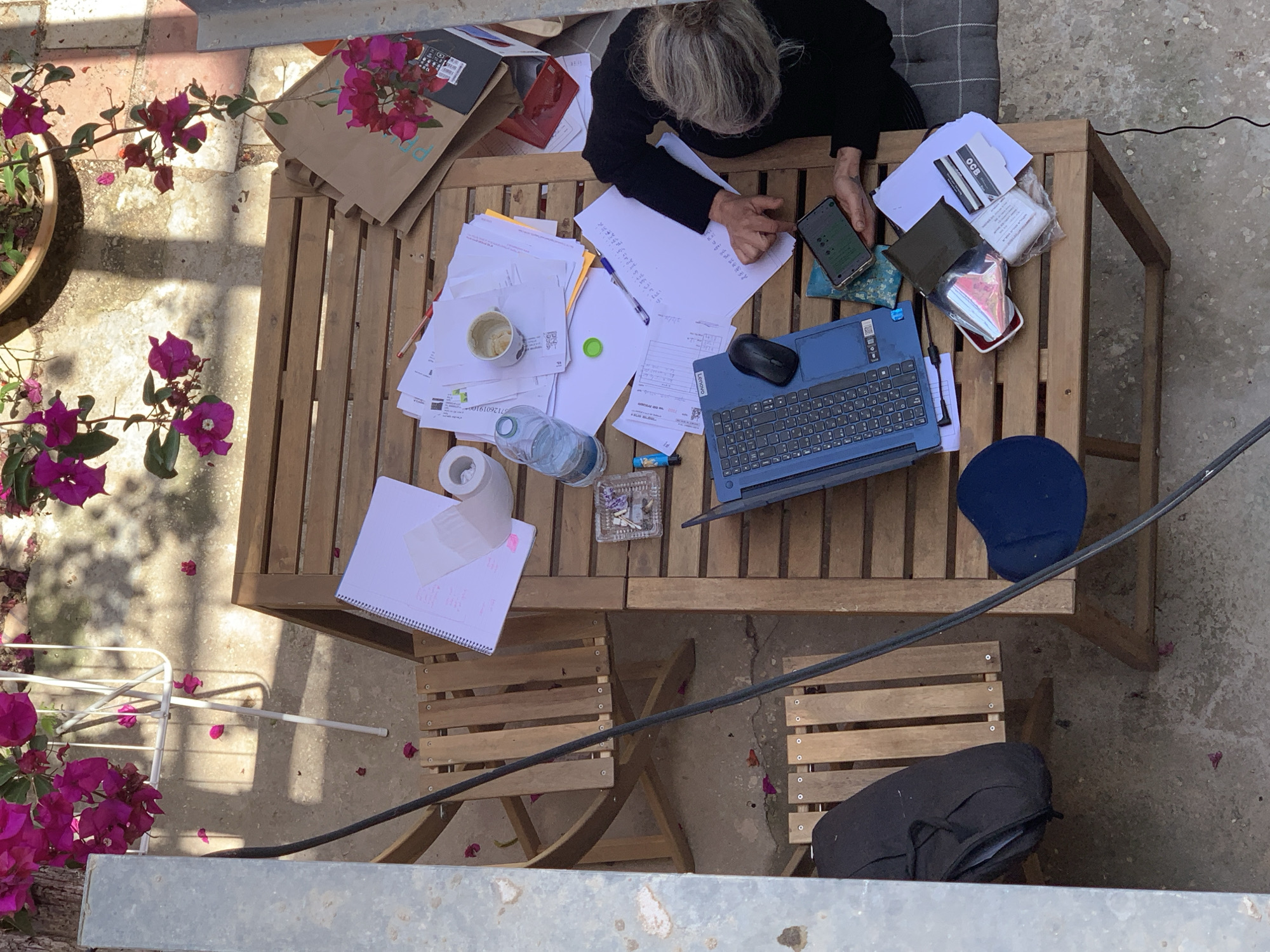 An overhead photograph captures a person engrossed in work or study at an outdoor wooden table on a sunny day. The scene unfolds on a concrete patio or balcony, featuring a single person with light-colored hair, wearing a dark top, seated at the table. Their face is not visible, but they are holding a smartphone in their left hand while their right hand interacts with documents on the table.
The wooden table is laden with numerous items, suggesting an active workspace. These include multiple stacks of papers with both handwritten notes and printed text, a blue Lenovo laptop equipped with a Hebrew keyboard, and an external black mouse. A ceramic mug, a plastic water bottle, a pen, and a roll of white tape are also scattered among the documents. A small, clear glass ashtray holds cigarette butts. Other personal items on the table include a brown paper bag with pink flowers and the text "ppdd", a red wallet, a small blue patterned pouch, and a small white plastic bag containing white items. An "OCB" branded card is visible on one stack of papers.
The immediate surroundings feature vibrant pink/purple bougainvillea plants in pots on the left, with fallen petals scattered across the concrete ground. Strong, sharp shadows cast by the table and an overhead structure indicate bright daylight and sunny weather. A blue mouse pad rests on the adjacent chair to the right, and a black backpack is placed on the chair below the table. The Hebrew keyboard on the laptop and the prominent bougainvillea plants offer subtle cues to the Tel-Aviv, Israel location.