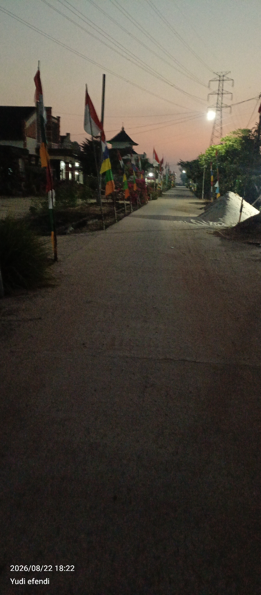 This is a vertical, low-light, first-person perspective image taken outdoors. The scene depicts a road with numerous Indonesian flags lining both sides, suggesting a celebration or national event. The flags are of various colors, including red and white, green, yellow, and blue. The road appears to be paved and leads into the distance. On the left side of the road, there are buildings and vegetation. On the right, there is a large pile of sand and more vegetation. In the background, a tall electrical tower with power lines is visible against a dusky sky with hues of orange and gray. The timestamp at the bottom left corner indicates the photo was taken on 2026/08/22 at 18:22 by Yudi efendi. The location is identified as Rawajitu Selatan, Indonesia. The overall atmosphere is subdued, likely due to the time of day.