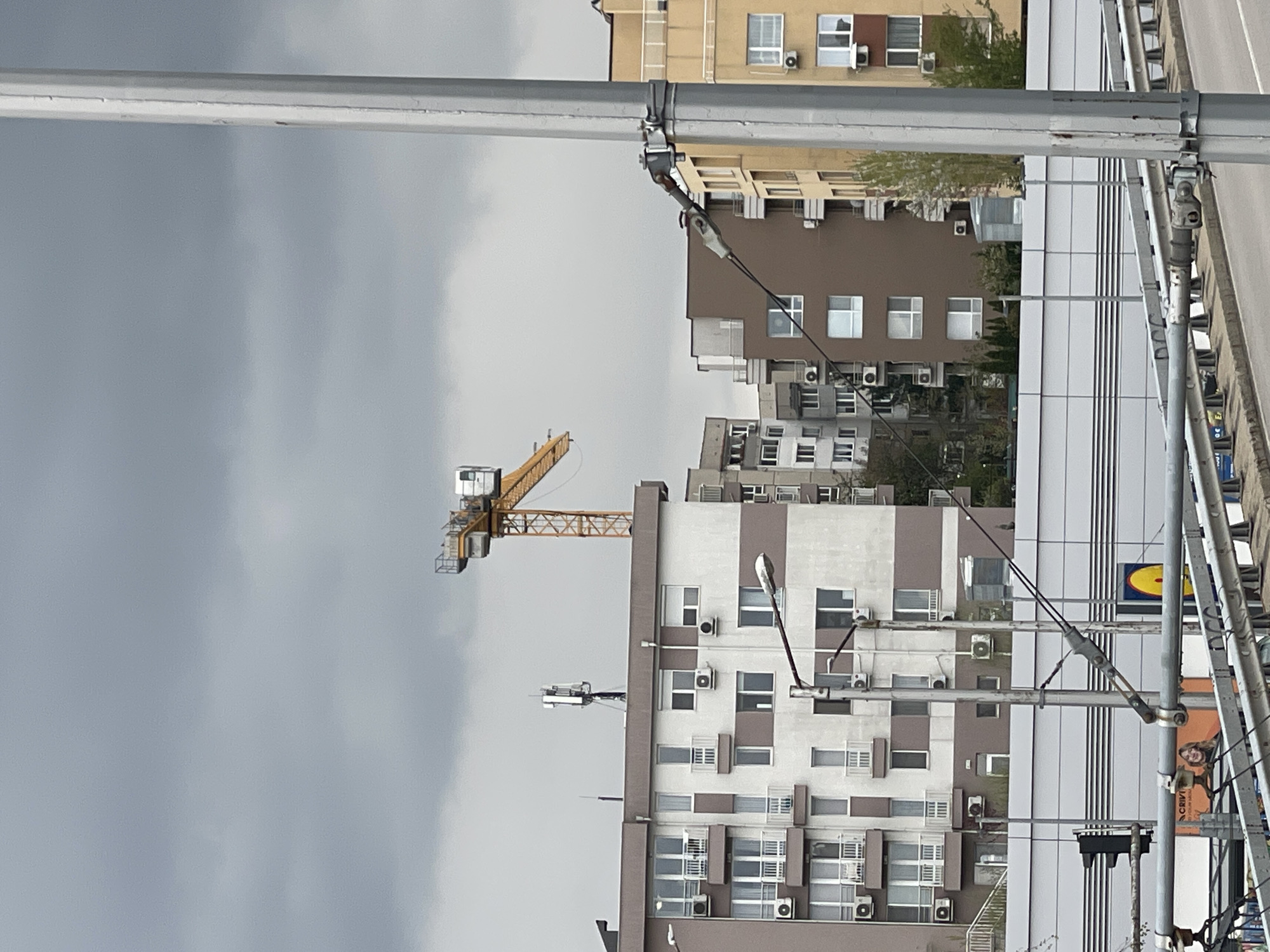 The image shows a cityscape under an overcast sky, likely in Sofia, Bulgaria, judging by the architectural style and the "LIDL" sign visible on a building. In the foreground, metal utility poles and overhead wires dominate the view, obscuring parts of the buildings behind. A yellow construction crane is visible to the left of a tall building with a white and brown facade punctuated by numerous windows and air conditioning units. Other buildings of varying heights and colors, including tan and brown structures with similar window and air conditioning unit arrangements, are present in the midground. A road with parked cars can be seen to the right of the building. There are no people visible in the scene.