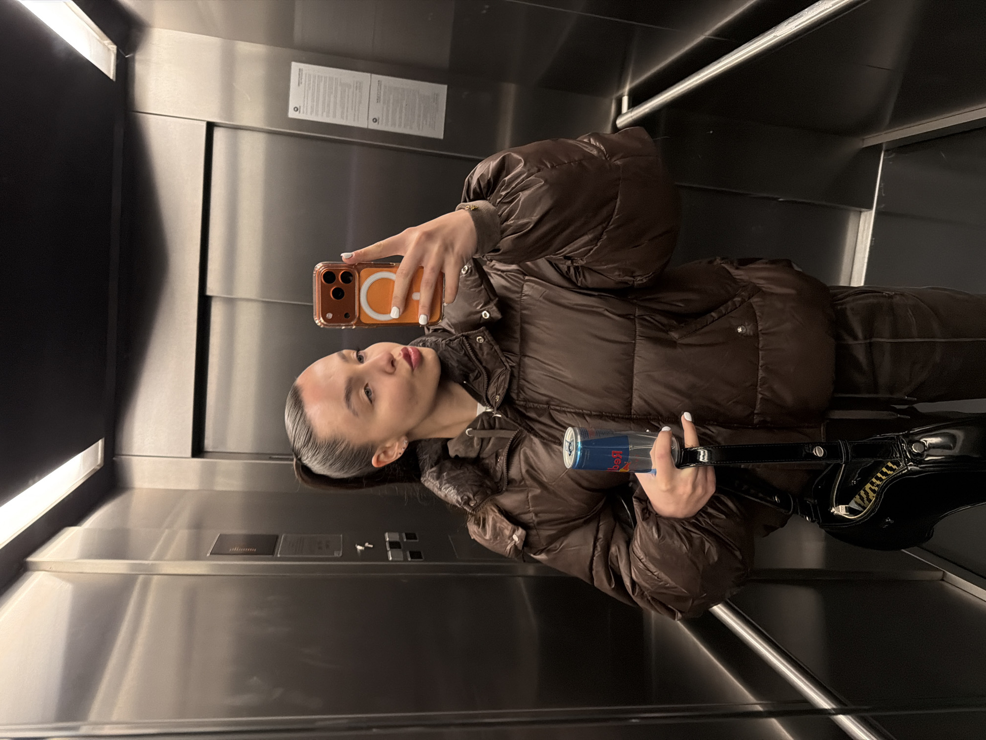 A young woman stands in an elevator, using her smartphone to take a picture of herself. She is wearing a brown puffer jacket. In her other hand, she is holding a can of Red Bull and a black purse. The elevator's interior is stainless steel, with a light fixture on the ceiling. A sign is visible above the elevator doors, and a metal bar runs along the side of the elevator car. The precise location in Sofia, Bulgaria, is not identifiable from the image.
