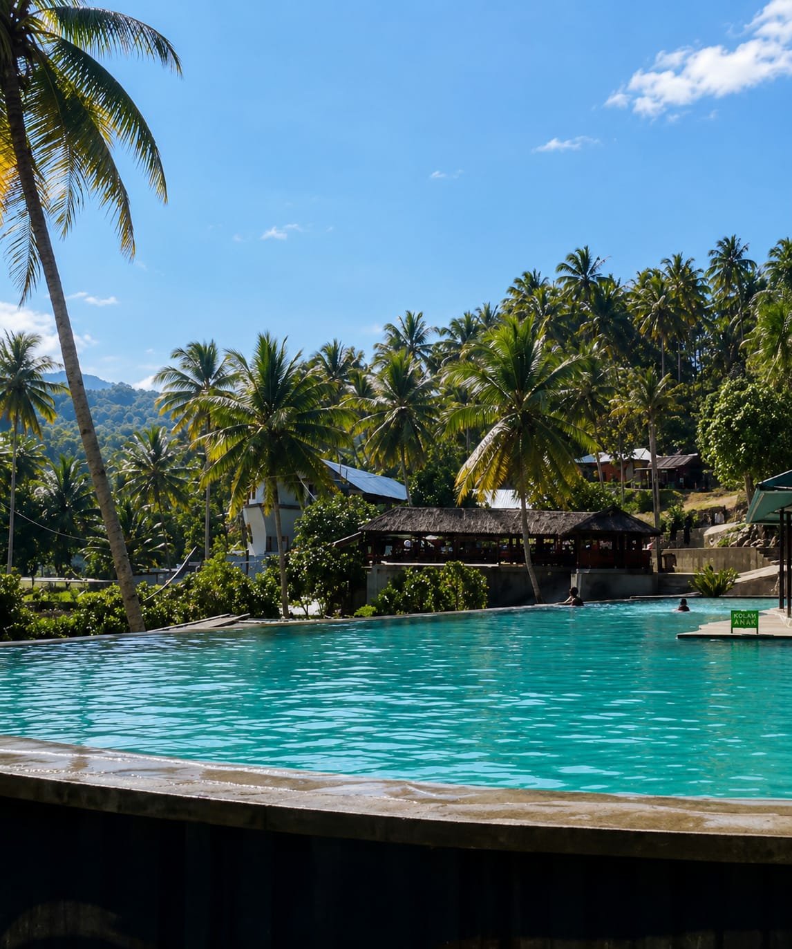 In Silaiyajae, Indonesia, a bright, clear day illuminates a luxurious infinity pool situated amidst lush tropical vegetation. The pool's turquoise water is calm, reflecting the vibrant blue sky dotted with wispy white clouds. Several palm trees, their fronds swaying gently, frame the scene, with one particularly large palm on the left casting a long shadow.
In the background, a cluster of buildings with thatched roofs and more modern structures are nestled among the trees, suggesting a resort or recreational area. Some of these buildings appear to be open-air structures, possibly restaurants or relaxation areas. A gentle slope of verdant hillside rises behind the buildings, further enhancing the tropical ambiance.
A few people are visible in the pool, their bodies partially submerged in the refreshing water, indicating a leisurely atmosphere. One person is swimming in the main pool, while another appears to be in a separate, shallower section marked with a sign that reads "KOLAM ANAK" (Children's Pool). The foreground of the image is dominated by the edge of the infinity pool, a concrete structure that creates a sleek, modern contrast with the natural surroundings. The overall impression is one of tranquility and relaxation in a beautiful, tropical setting.