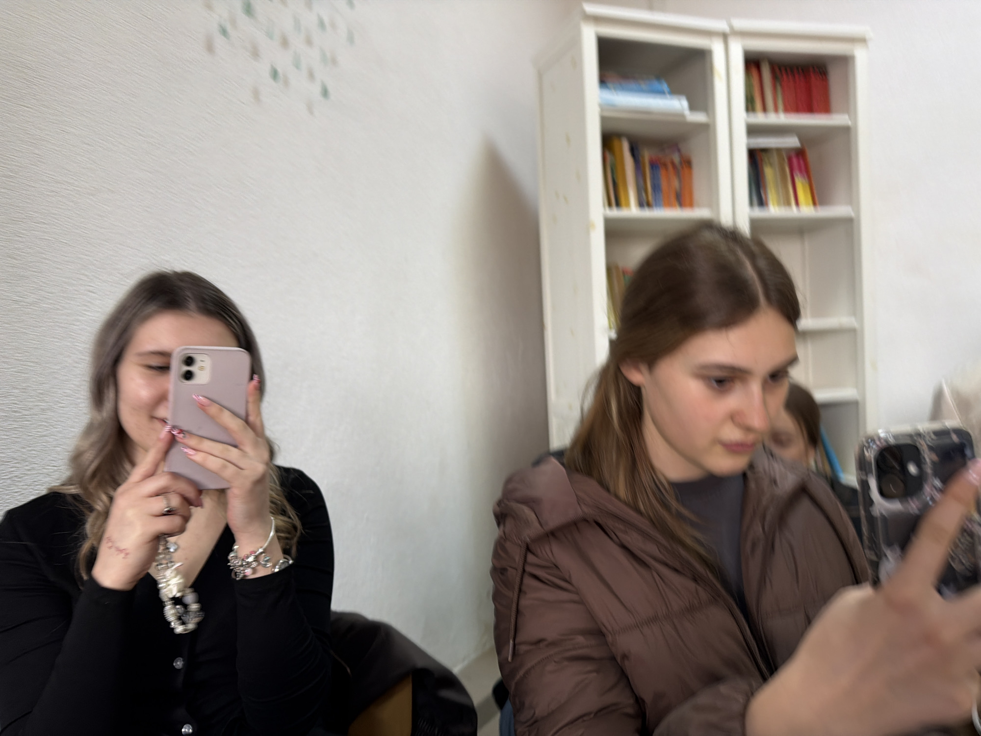 Two young women are the primary subjects in the scene. The woman on the left is taking a picture of herself, using a pink smartphone with two camera lenses, wearing a black buttoned shirt with long sleeves, and jewelry including bracelets and a necklace. The woman on the right is also holding a smartphone, possibly taking a picture as well, wearing a brown puffer jacket. Between the two women, in the background, is a white bookcase filled with books, having red and yellow spines. The background wall is painted white. There is also a wall decoration with small colored shapes, also in the background. The location is inside, presumably in the city of Panagyurishte, Bulgaria.