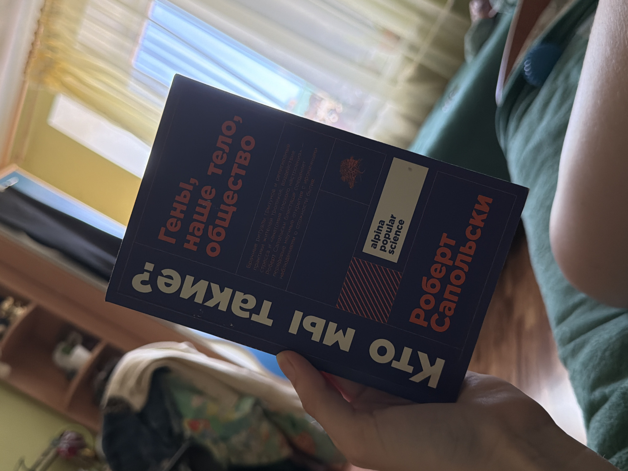 A close-up, eye-level, slightly upward-angled shot shows a hand holding an open book. The book's cover is dark blue with bold, white, and orange text. The title reads, "КТО МЫ ТАКИЕ?" (Who are we?) in large white letters oriented vertically on the left side of the cover. To the right, in orange, it says "ГЕНЫ, НАШЕ ТЕЛО, ОБЩЕСТВО" (Genes, Our Body, Society). Below this, there is smaller text and a logo for "alpina popular science". At the bottom of the cover, the author's name, "Роберт Сапольски" (Robert Sapolsky) is printed in orange. The background is blurred, with a wooden shelf unit on the left and a window with yellow curtains on the right, suggesting an indoor environment, possibly a home or office. The lighting appears to be natural, coming from the window. The image does not contain any identifiable landmarks or elements that would place it specifically in Zavodoukovsk, Russia.