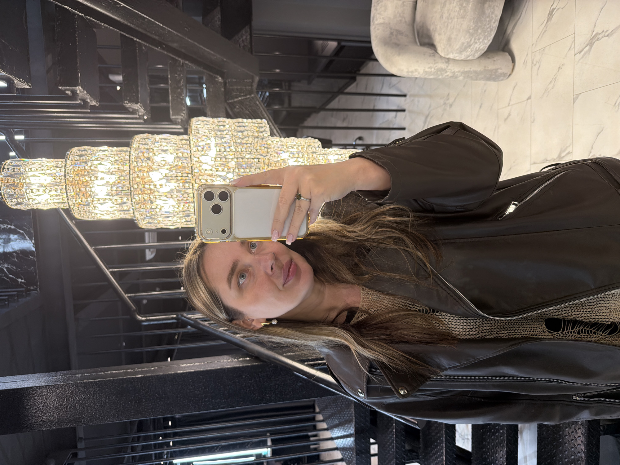 The image displays a woman taking a selfie in a luxurious indoor setting, identified as being in Moscow, Russia. The photograph is oriented with the woman's head tilted sideways, implying it was originally taken in portrait mode and then rotated 90 degrees counter-clockwise.
The main subject is a woman with long, wavy light brown hair featuring blonde highlights. She looks towards the camera with light eyes and a calm, slight smile. She is dressed in a dark brown leather jacket worn over a light-colored, possibly beige, textured top. Notable accessories include a ring with a dark stone on her left ring finger and small gold earrings. She is holding a smartphone, identifiable as an iPhone by its camera module, encased in a light-colored cover with a gold or silver trim, using it to capture the selfie.
The background reveals a sophisticated interior space. To her left, a grand, multi-tiered chandelier composed of numerous sparkling crystals illuminates the scene with a warm glow. Behind her and extending into the distance is a modern staircase with dark metal railings and textured steps, suggesting a multi-story structure. Further background elements include a dark, possibly black marble surface to the left and a light-colored marble-patterned floor or wall on the right, where a blurry, abstract light-colored sculpture or furniture piece is also visible.
The overall setting suggests an upscale environment, such as a hotel, a high-end residence, or a luxury commercial space, characterized by contemporary design, opulent lighting, and premium materials. No discernible text is visible in the image. The time of day is not evident, as the lighting primarily comes from artificial sources.