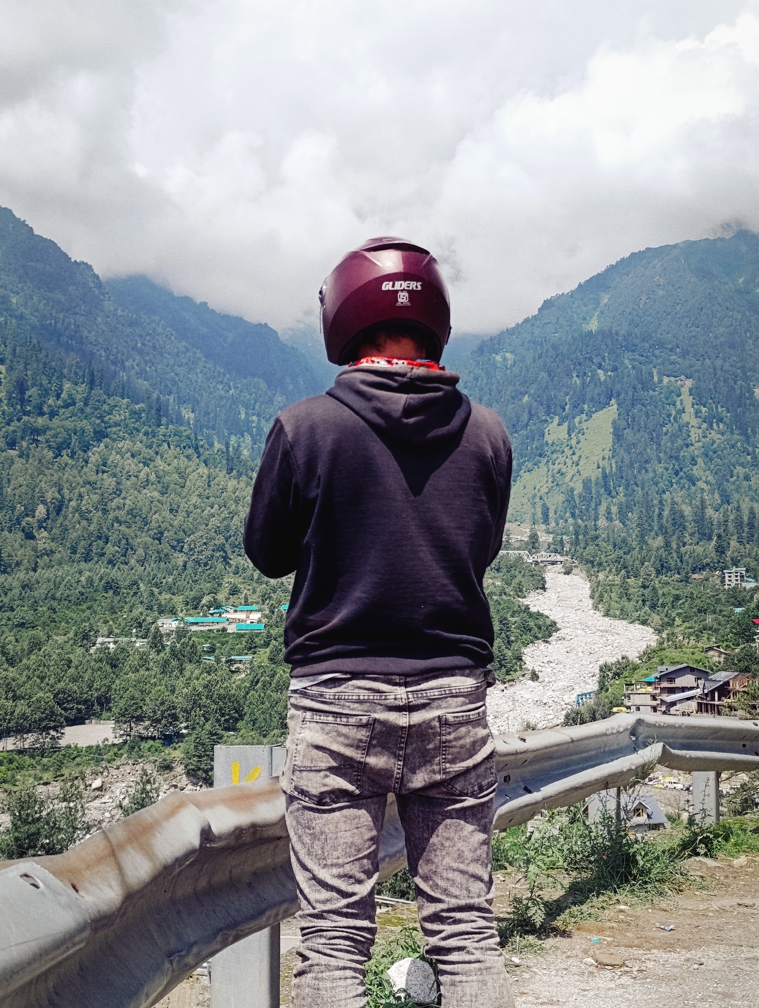 A person wearing a maroon helmet and a black hooded sweatshirt stands with their back to the camera, looking out at a mountainous landscape. The person is wearing grey jeans. The landscape features lush green trees covering the slopes of large mountains, with a cloudy sky above. A riverbed filled with rocks runs through the valley, and a few buildings are visible in the distance, nestled amongst the trees. A guardrail runs along the foreground. The image appears to be taken in Palchan, India.