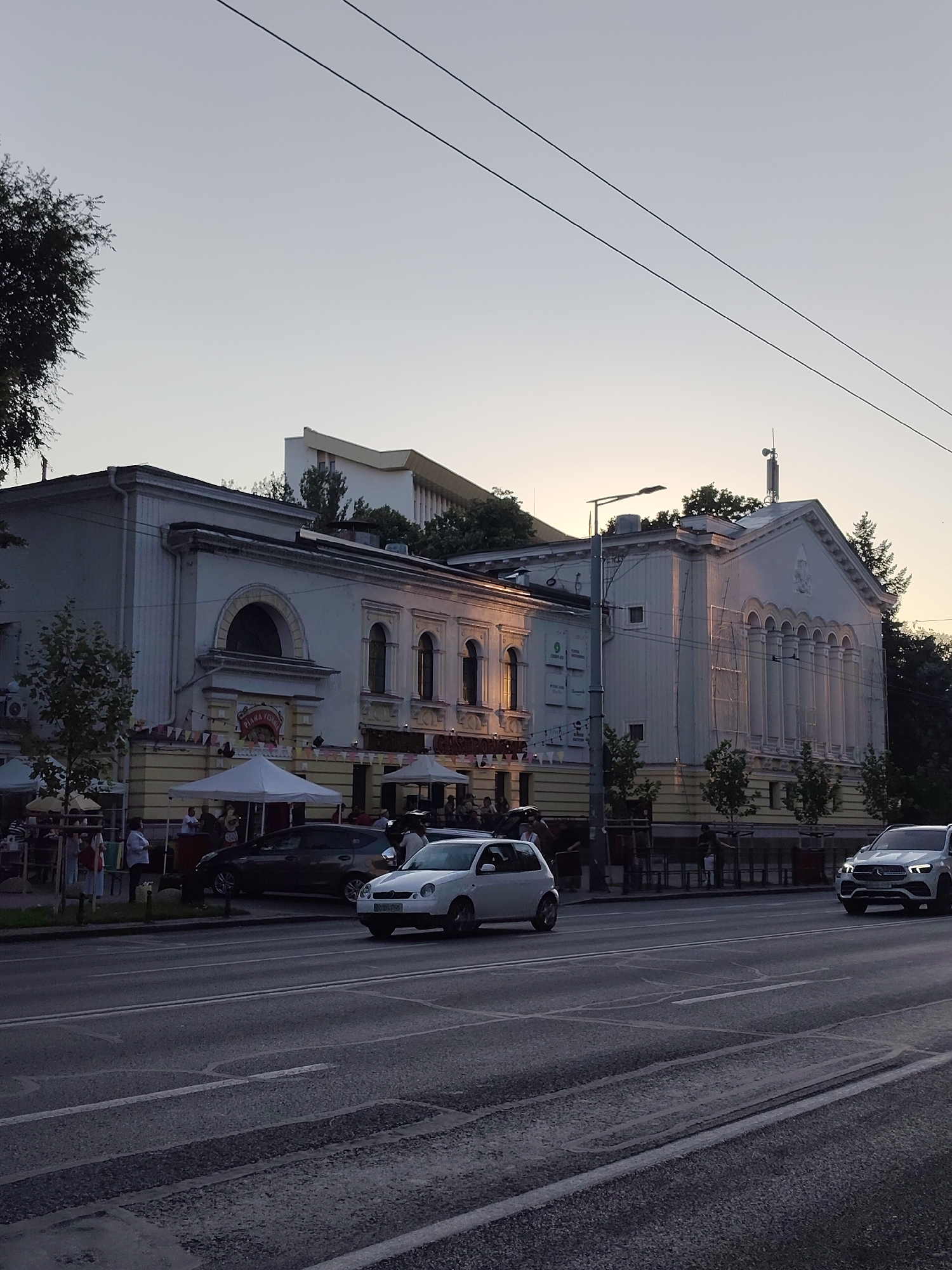 In Chișinău, Moldova, a street scene unfolds at dusk with a white Mercedes-Benz SUV in the foreground, moving right, and a white compact car in the middle ground, also heading right. Behind them, an older white building with ornate architectural details and arched windows lines the street. The building appears to be a performance venue or cultural center, with banners and awnings suggesting an outdoor seating area or event. Several cars are parked along the street, and people are visible walking and gathered under tents in front of the building. Overhead, power lines crisscross the sky, and the light suggests a late afternoon or early evening setting.