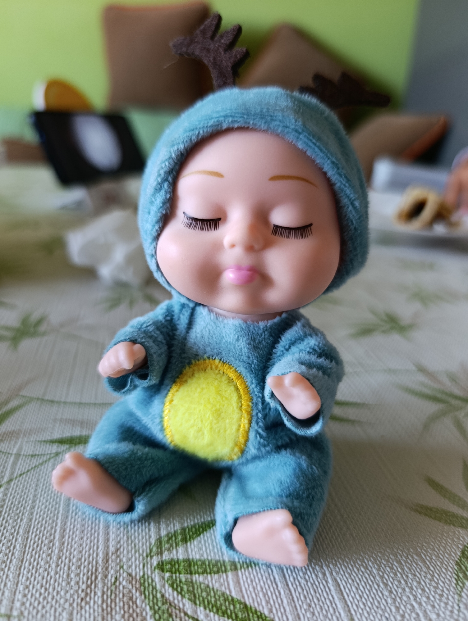 A close-up shot shows a small, blue doll with a yellow patch on its chest sitting on a patterned surface. The doll has a light skin tone, closed eyes with pronounced eyelashes, and small brown antlers attached to its hood. The background is blurred, but includes hints of a green surface, pillows, and possibly a tablet. The location in Plovdiv, Bulgaria is not apparent from the image.