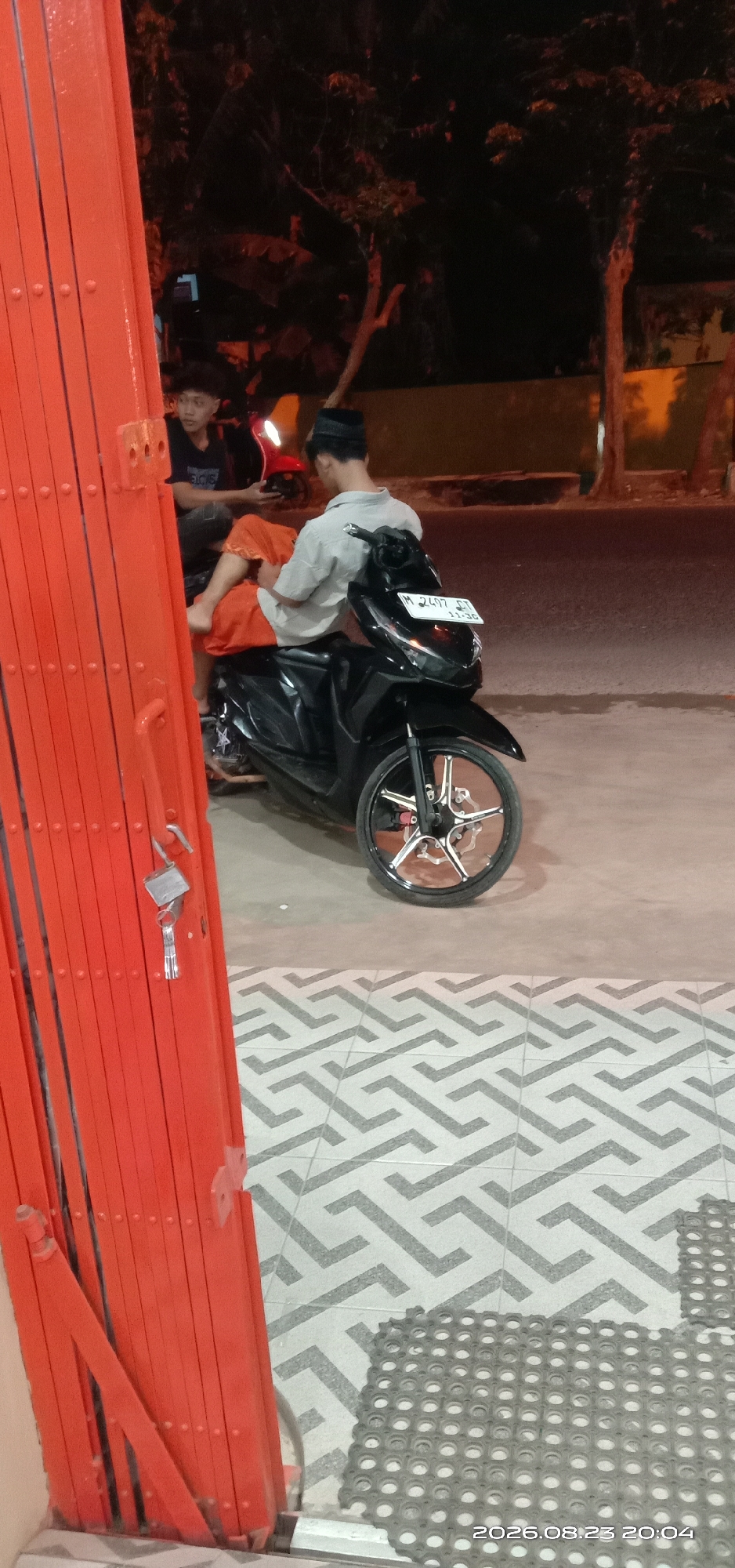 In the city of Pakong, Indonesia, two young men are observed outdoors at night, near an orange retractable gate. The man on the left, wearing a black t-shirt, is seated on a red motorcycle, looking towards his companion. The man on the right, seated on a black motorcycle, wears a light-colored shirt and orange shorts and has a black cap on his head. His focus is directed downwards. The motorcycles appear to be parked on a tiled surface with a geometric pattern. A lock and keys are attached to the orange gate in the foreground. The background is dark and features trees and street lighting. The timestamp on the image indicates August 23, 2026, at 20:04.