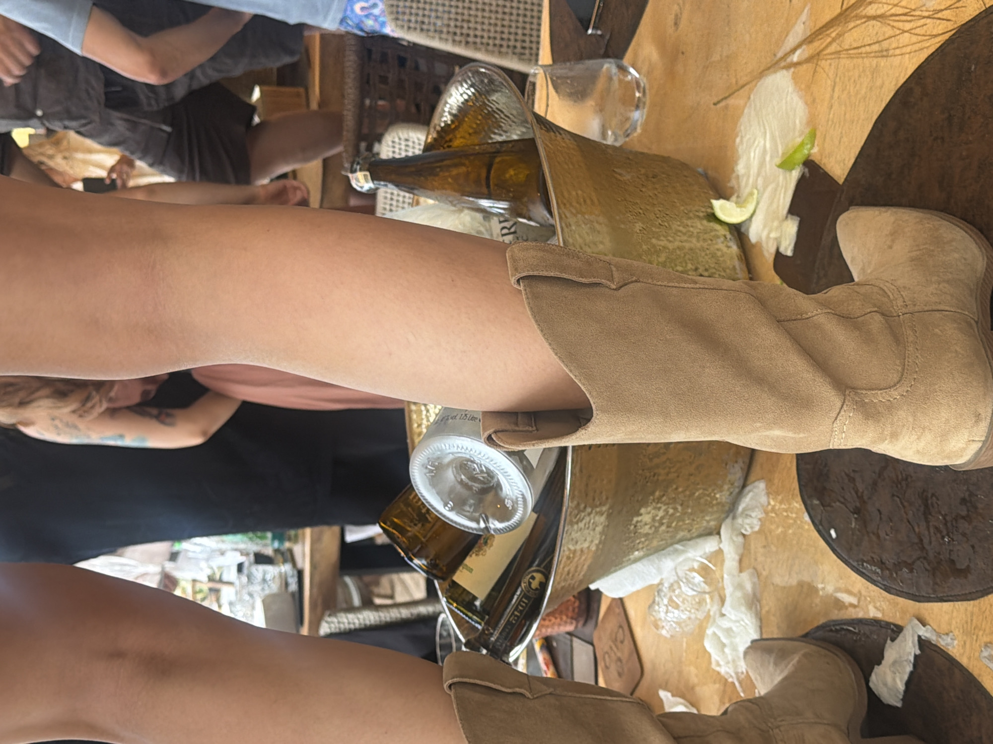 An overhead shot captures a portion of two individuals standing in what appears to be an outdoor dining or bar area, possibly at a resort. The foreground features the lower legs and feet of two people, both wearing tan-colored cowboy boots, positioned over dark, circular placemats that are wet. The left boot is more prominent, with its leg partially obscuring the view of the other.
Between the two sets of legs, a large, hammered metallic gold ice bucket is centrally placed on a wooden table. The bucket is filled with several empty brown glass bottles, one white bottle, and some crumpled white napkins or paper. A few pieces of lime and some white residue are scattered on the table around the ice bucket, suggesting a recent serving of drinks.
In the background, other individuals can be seen, some with visible tattoos on their arms, and the general ambiance suggests a casual social gathering. The lighting indicates it is daytime. The flooring or table surface is a warm-toned wood.