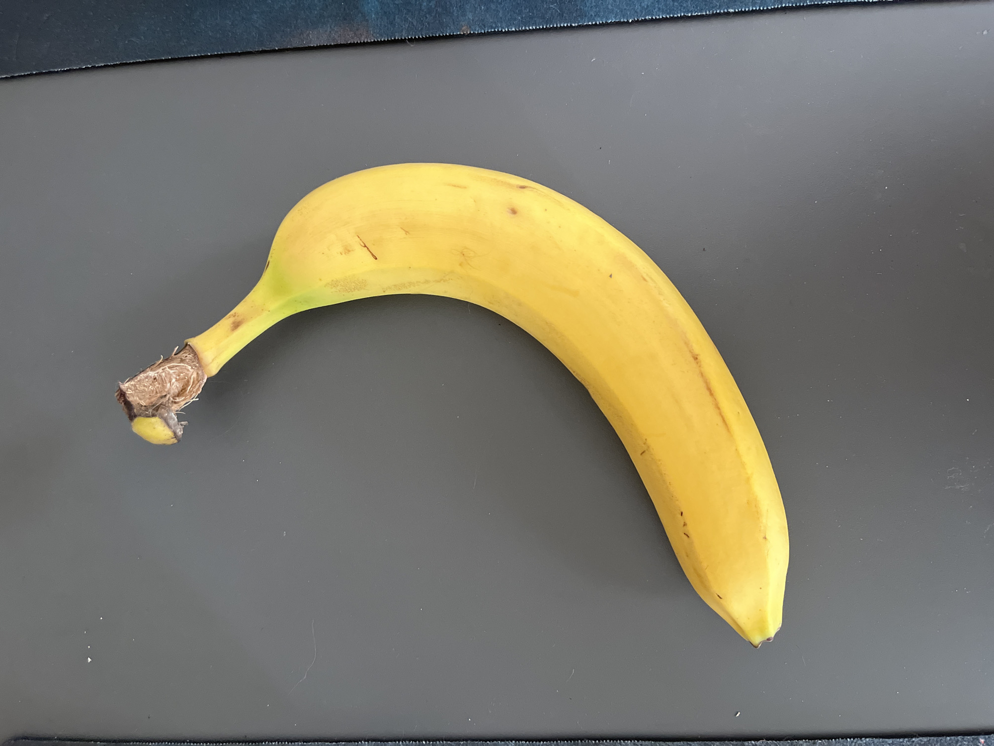 A single, ripe yellow banana is positioned diagonally on a dark gray, flat surface. The banana is slightly curved, with its stem end at the top left and the tip pointing towards the bottom right. The surface appears to be a desk or countertop, with some faint dust specks visible. The background on the right edge of the image shows a textured blue material. There are no people, actions, or interactions depicted. The location is not identifiable as Shymkent, Kazakhstan, or any other specific city.