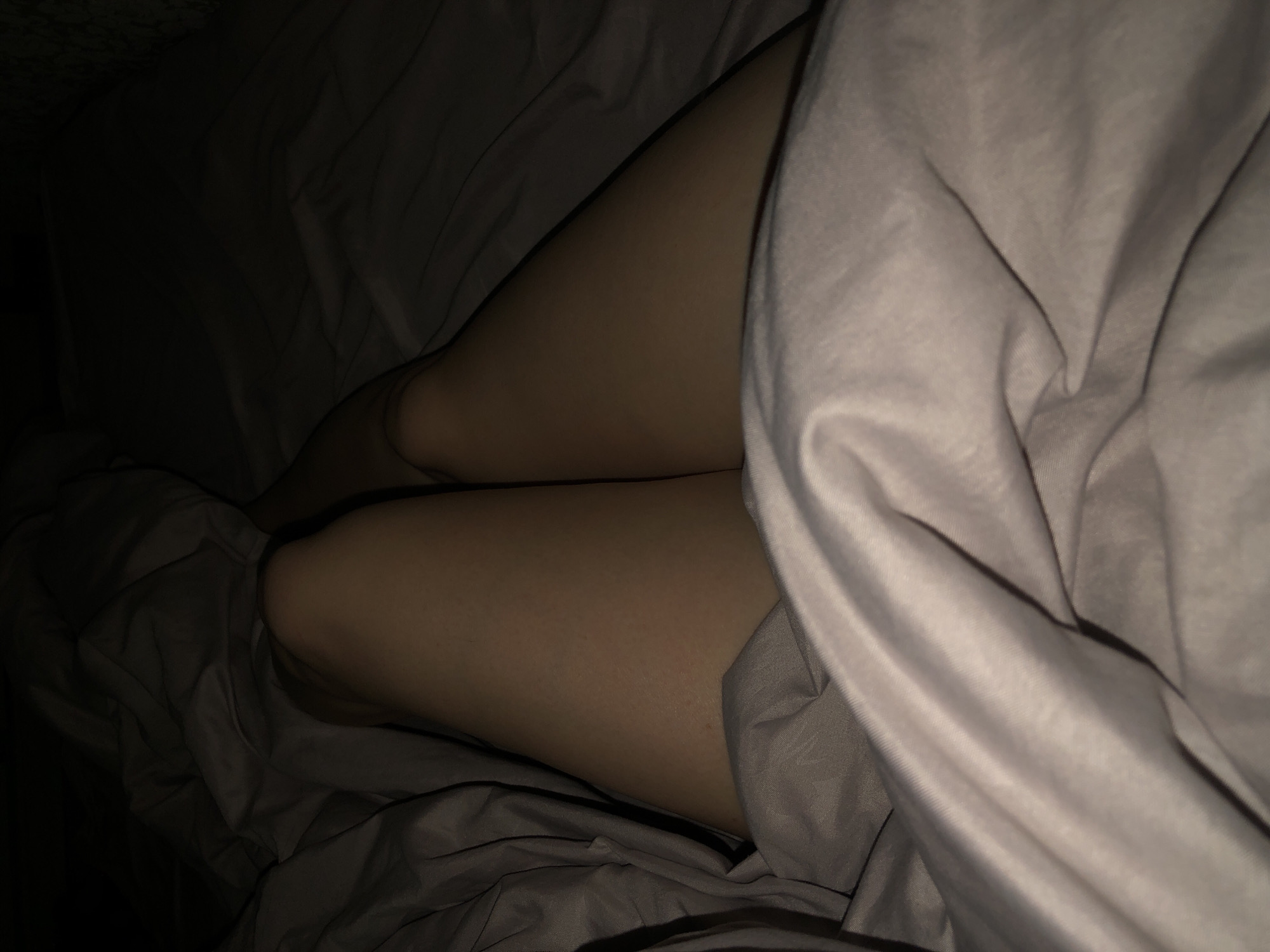 The image depicts a person's legs under a blanket in dim lighting. The person is lying down on a bed with gray sheets and blankets. The location is not specified in the image.