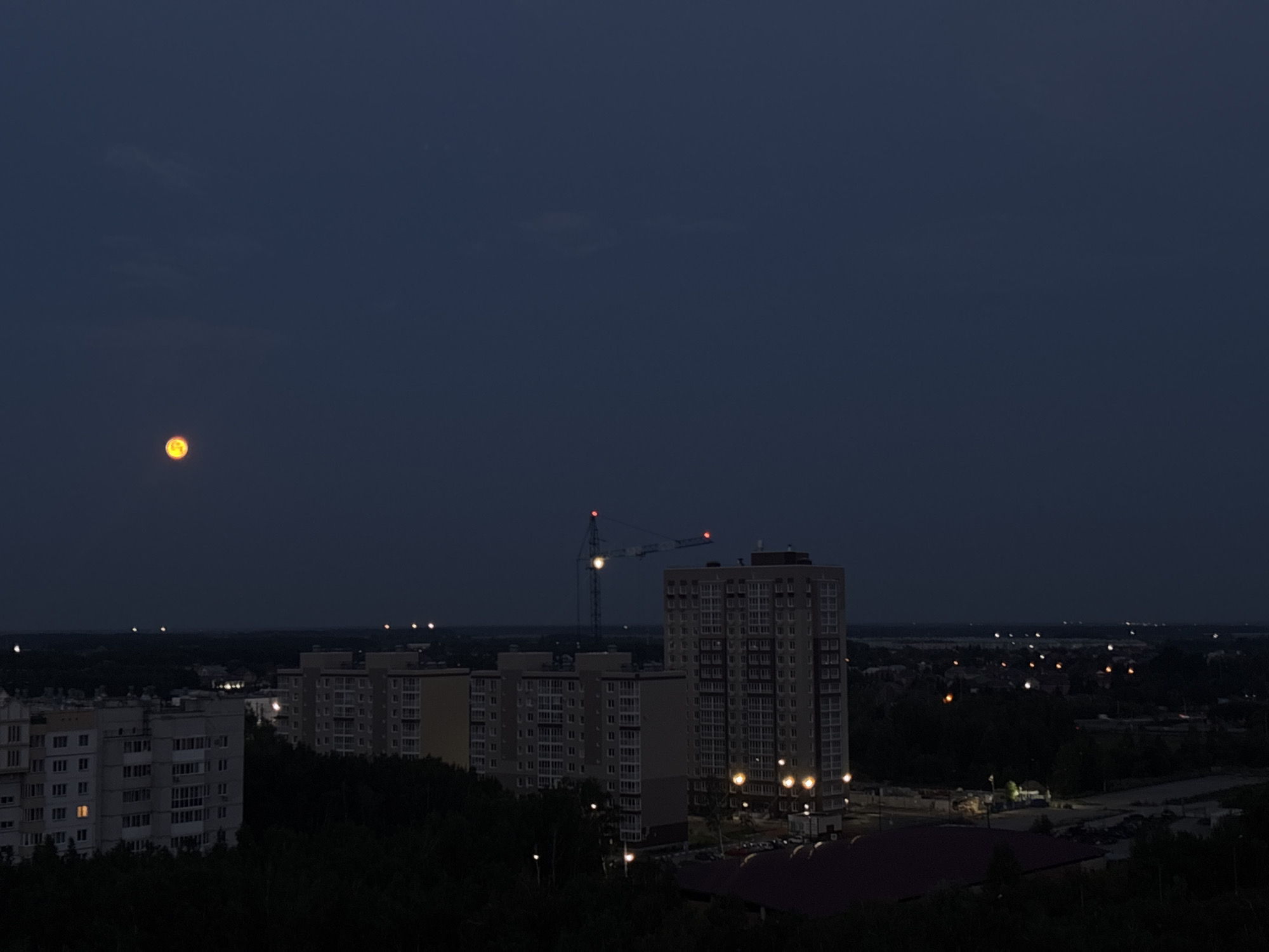 This is a night aerial view of a city, likely Omsk, Russia, with a dark blue sky and scattered lights indicating urban activity. In the upper left quadrant, a glowing orange object, possibly the moon, is visible. The cityscape features several tall apartment buildings, some with illuminated windows, and a construction crane with red lights at its apex, suggesting ongoing development. Rows of trees form dark silhouettes in the foreground and middle ground, bordering the built-up areas. Distant lights create a hazy horizon, further emphasizing the vastness of the urban expanse under the night sky.
