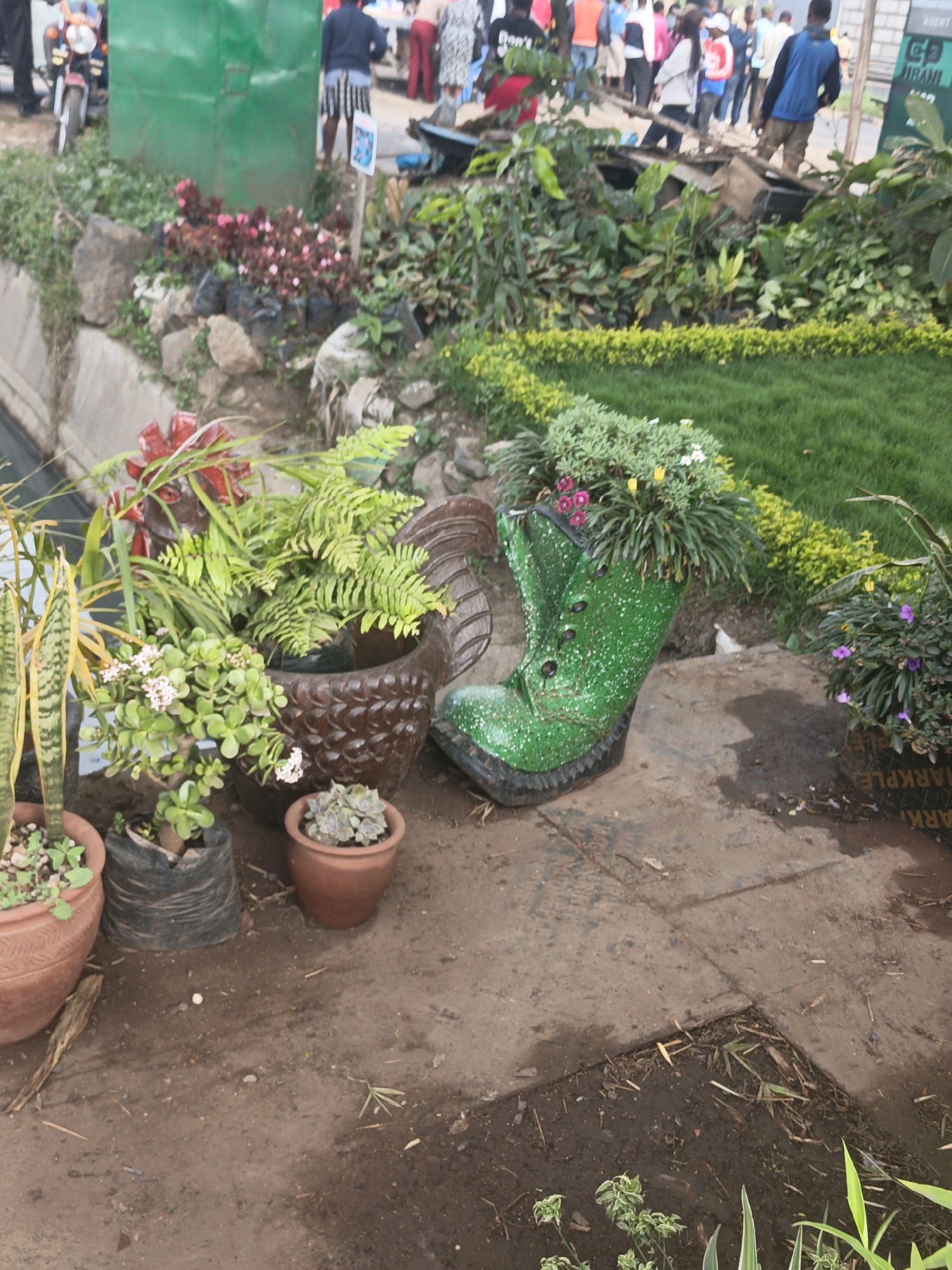 The image shows a roadside display of potted plants in Nairobi, Kenya. In the foreground, there are several flower pots of various sizes and materials. A large, green boot-shaped planter is filled with flowering plants. Adjacent to it is a dark brown, chicken-shaped planter overflowing with ferns and other greenery. Small pots with succulents and snake plants are scattered around. The ground is dark and damp. In the background, a crowd of people is visible, some walking and some standing. A green metal shed, a motorcycle, and various plants and flowers are also visible. The overall impression is that this is a makeshift plant stall or display alongside a busy street.