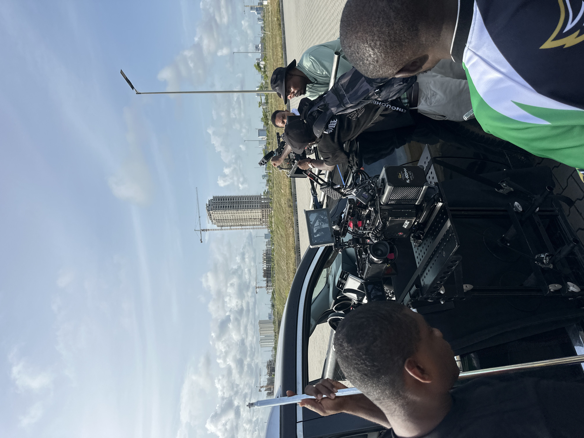 Several individuals are gathered around a black vehicle, likely involved in a film or video production. The vehicle is equipped with an extensive camera setup, including cameras, monitors, and support rigging. People are focused on the equipment and appear to be adjusting or operating it. In the background, construction is visible, with buildings and cranes indicating ongoing urban development. This scene appears to be taking place in Lagos, Nigeria.