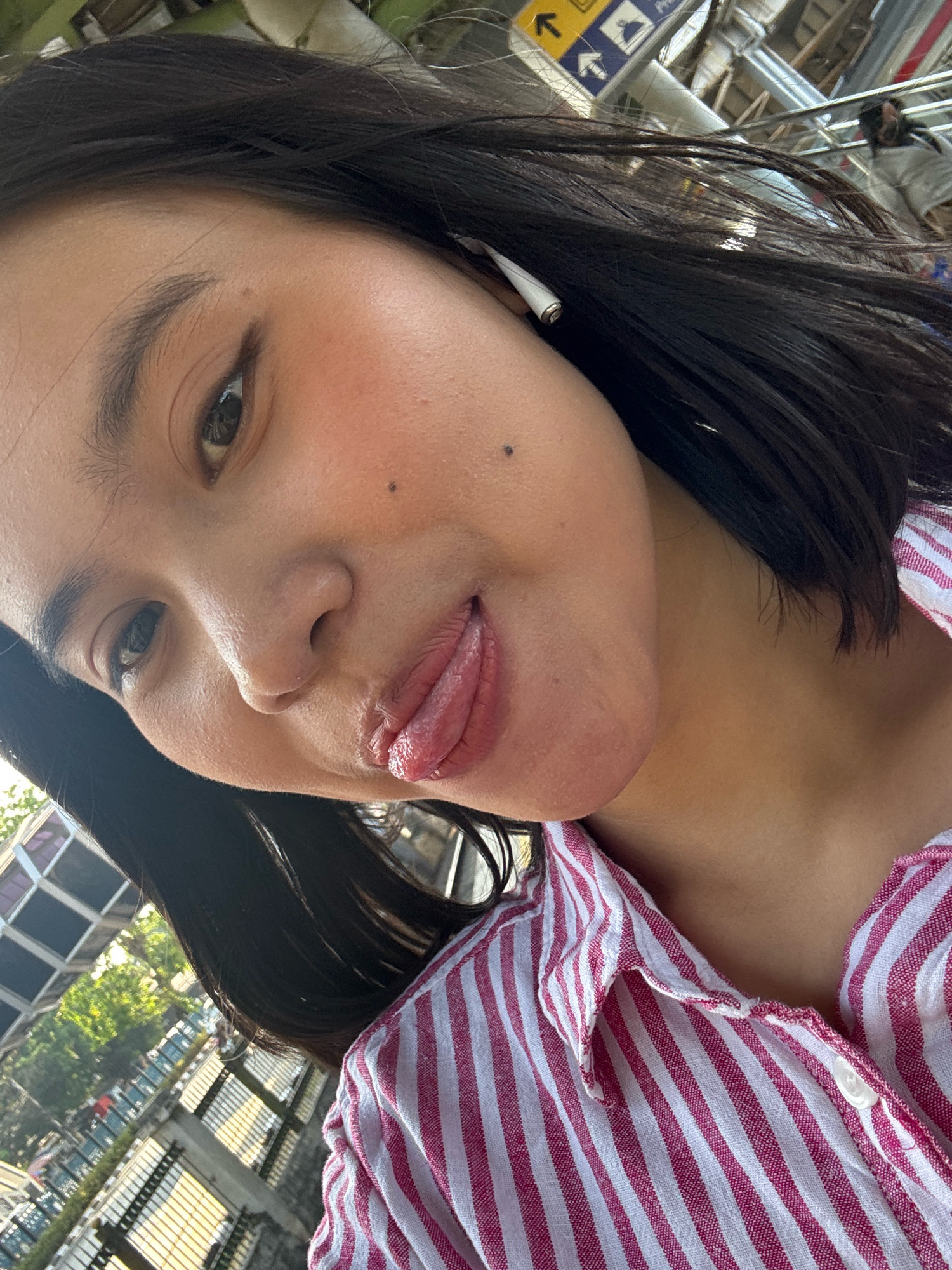 A young woman with dark hair and an earbud in her right ear is taking a selfie. She is wearing a pink and white striped shirt. Her tongue is sticking out slightly. The background appears to be an outdoor public space with some buildings and railings visible, suggesting it could be a transit station or a pedestrian walkway in a city. The lighting indicates it is daytime. The overall scene suggests a casual and candid moment.