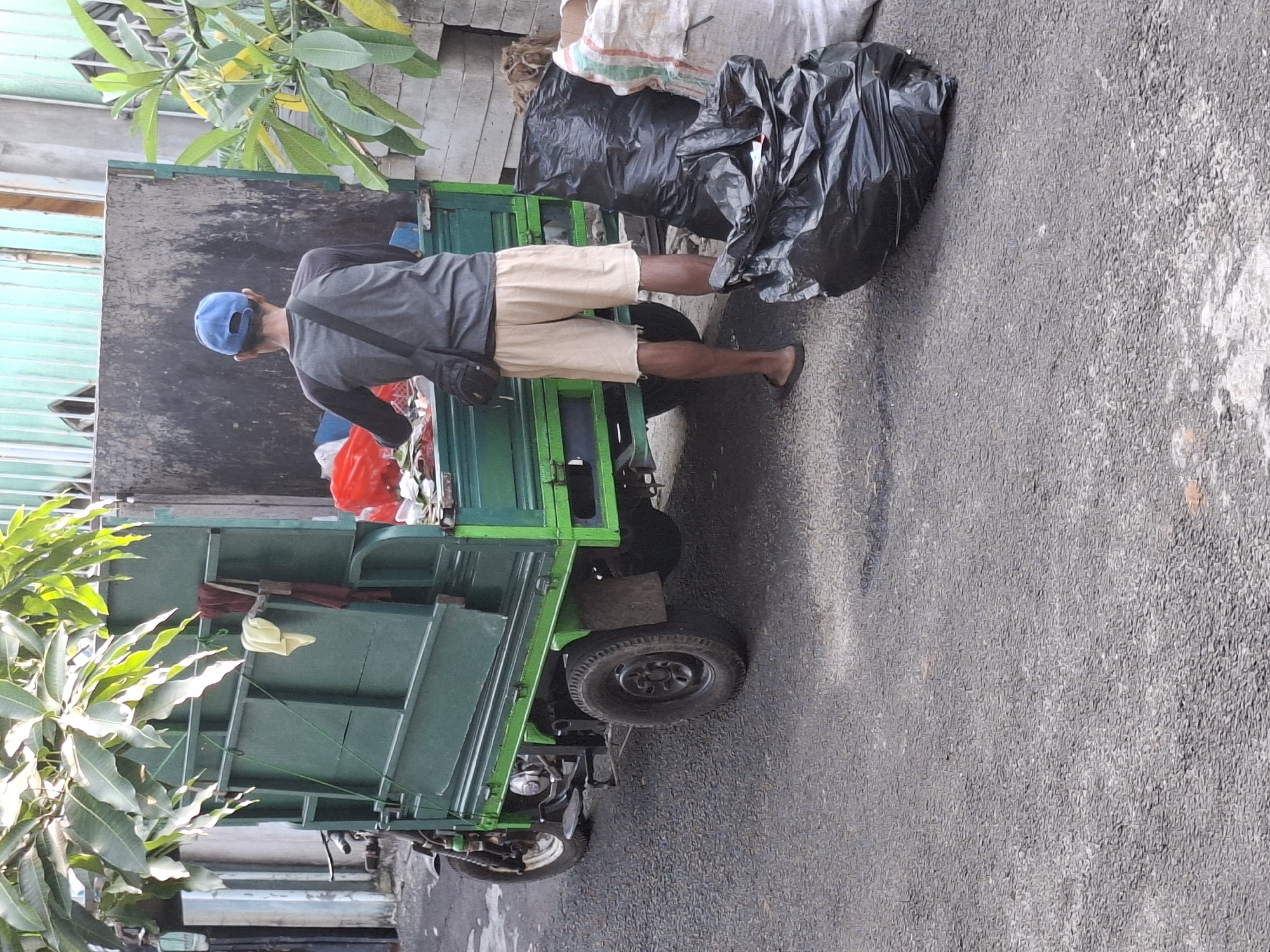A person wearing a blue cap, gray long-sleeved shirt, tan shorts, and black flip-flops is standing next to a green garbage truck in West Jakarta, Indonesia. The person is facing away from the camera, looking into the open back of the truck, which is filled with various items, including red plastic bags and white debris. Two large black garbage bags are on the ground to the right of the truck, and a lighter colored woven bag is next to them. The truck itself appears to be a three-wheeled vehicle with a large cargo bed. The ground is paved with asphalt. Lush green foliage from trees hangs over the truck from the left side of the image. In the background, there is a pale blue and white wall and a green metal fence or gate.