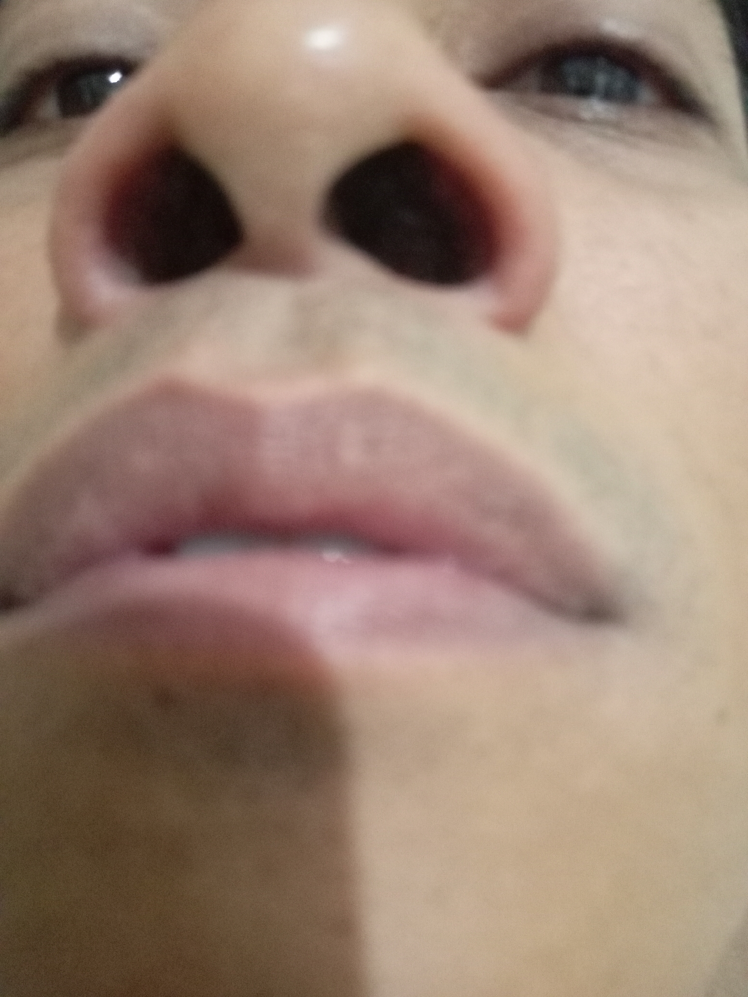 The image is a close-up, low-angle shot of a person's face, focusing on the nose and mouth area. The lighting is harsh, creating strong shadows, particularly on the left side of the face. The image is blurry and lacks fine detail, suggesting it may have been taken with a low-resolution camera or in poor lighting conditions. There are no identifiable landmarks or context to place it in Batam, Indonesia, or any other specific location. The subject's skin tone appears to be light to medium. The eyes are partially visible in the upper corners of the frame, and the nostrils are dark and prominent. The lips are full and appear to be slightly parted. There is no discernible action or interaction taking place.