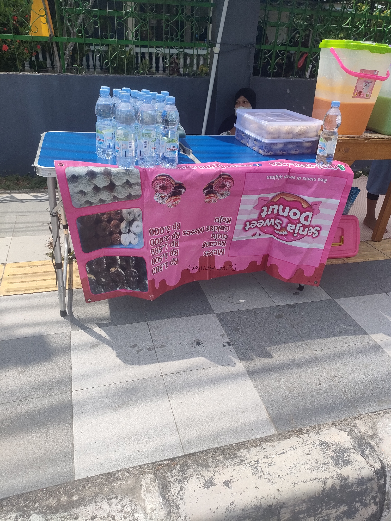 In Sungailiat, Indonesia, a street vendor displays an assortment of donuts and bottled water on a blue folding table. The donuts, offered in various flavors including chocolate messe, cheese, and sugar, are presented on a pink banner that also advertises "Senja Sweet Donut". A woman, partially visible in the background, is seated behind the table. Adjacent to the table, large beverage dispensers hold what appears to be iced tea or juice. The scene is set on a paved sidewalk, with a green fence and lush vegetation visible in the background. The sun casts long shadows, indicating it is likely mid-day.