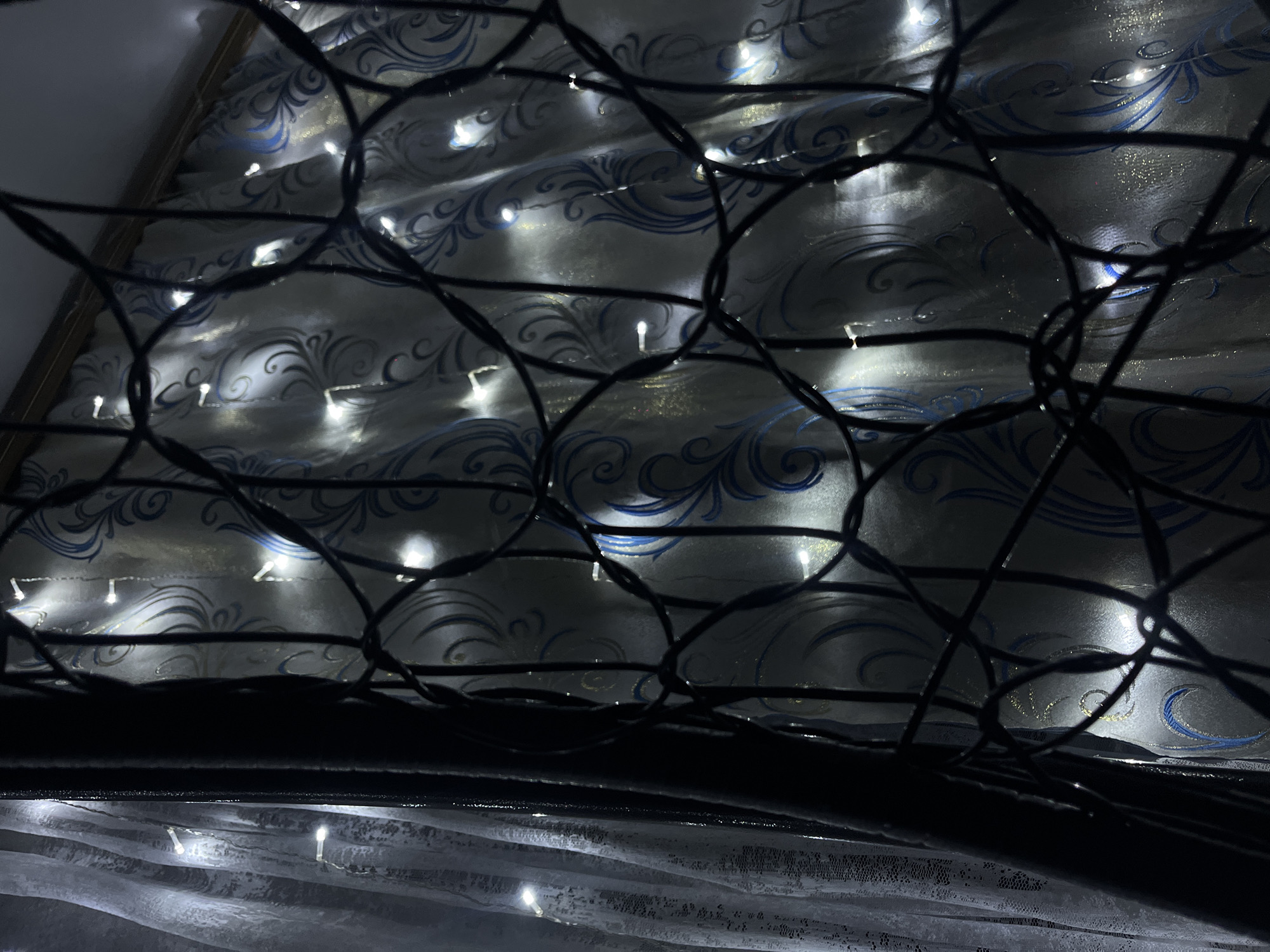The image shows a curtain with a silver and blue swirling pattern. String lights are interspersed throughout the curtain. A black net-like structure is in the foreground, partially obscuring the curtain and lights. The bottom of the image features a dark, possibly reflective surface, and a portion of a white, lacy curtain is visible beneath it. The specific location in Novopokrovskaya, Russia is not identifiable from the image. No people, specific subjects or actions are visible. Interactions cannot be determined. The environment is indoors.
